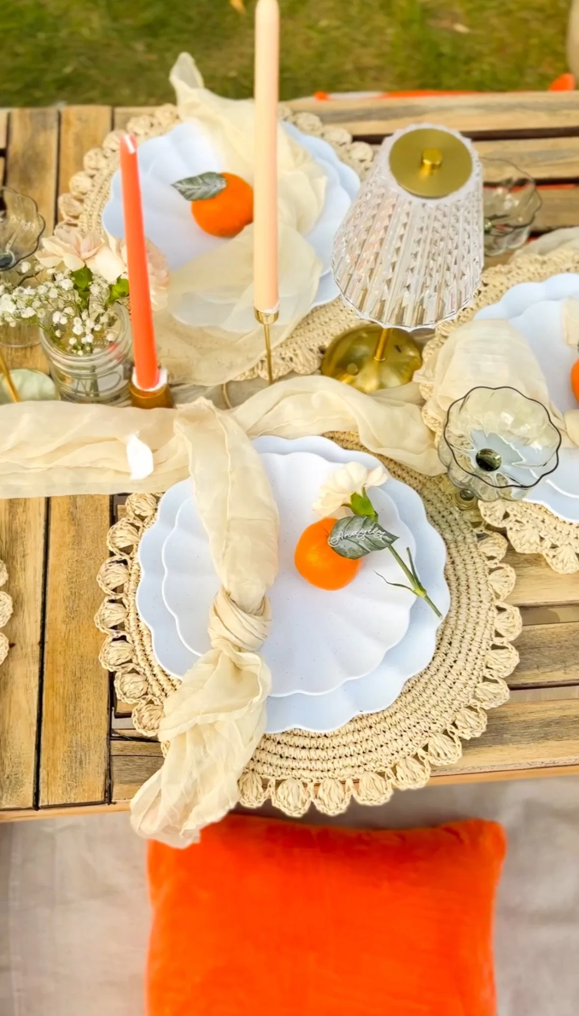 Decorative luxury picnic table setting with a white scalloped plate, orange fruit with a green leaf, pink and orange candles, a small floral arrangement, a lace-trimmed tablecloth, a table lamp, and a wooden table with an orange pillow.