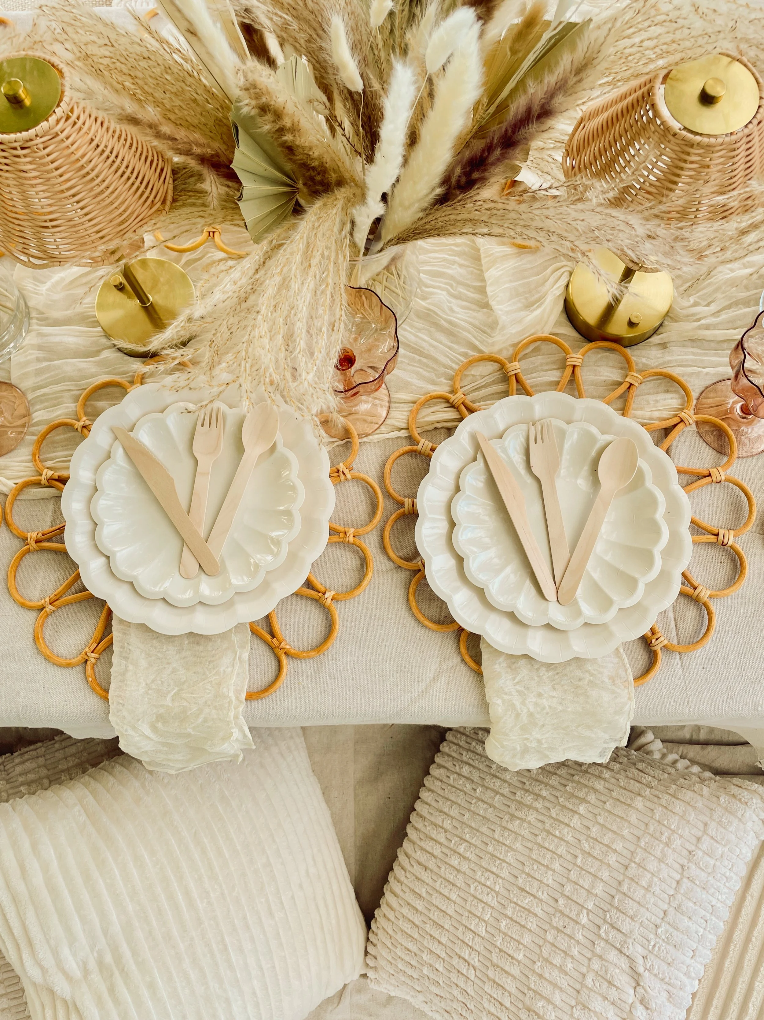A table setting featuring white plates, wooden cutlery, and decorative elements including tall dried pampas grass, green and amber glassware, and woven wicker vases, arranged on a beige tablecloth with a woven placemat underneath.