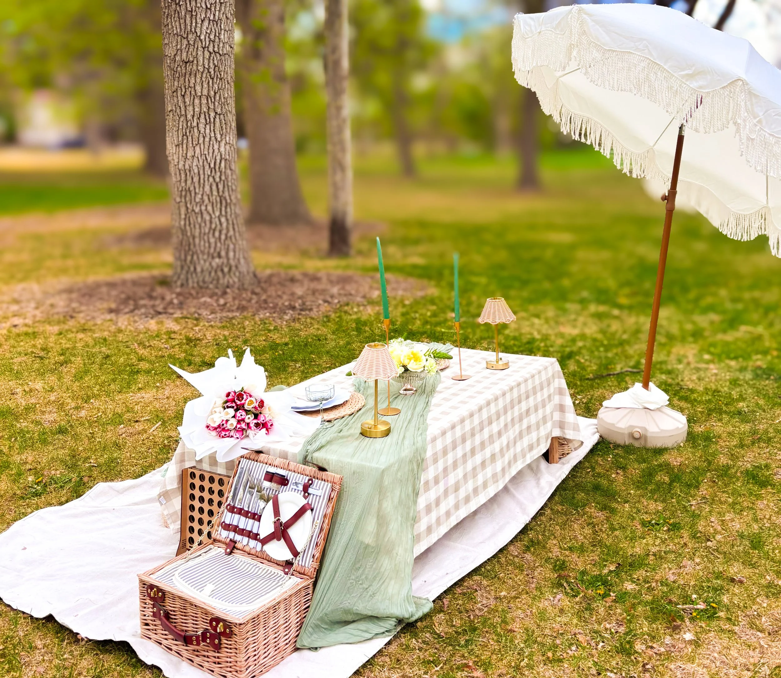 Outdoor picnic setup with a checkered tablecloth, flower arrangements, decorative umbrellas, and baskets, situated on grass with trees in the background.