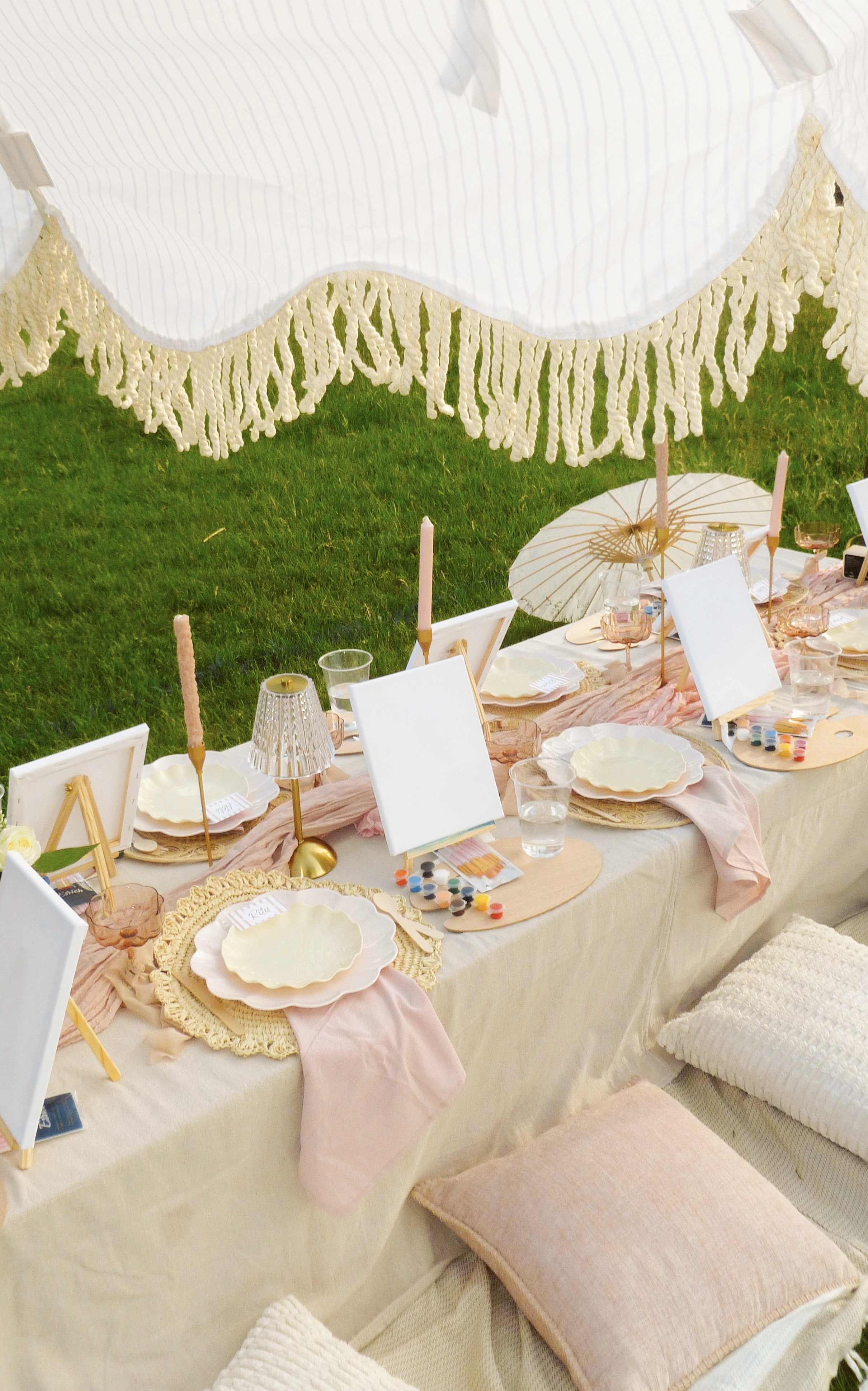 A decorated outdoor dining table set with white plates, pink napkins, candles, and small framed signs on beige tablecloth, with pink and white accents, under a white canopy with hanging knit fringe, on green grass patio.