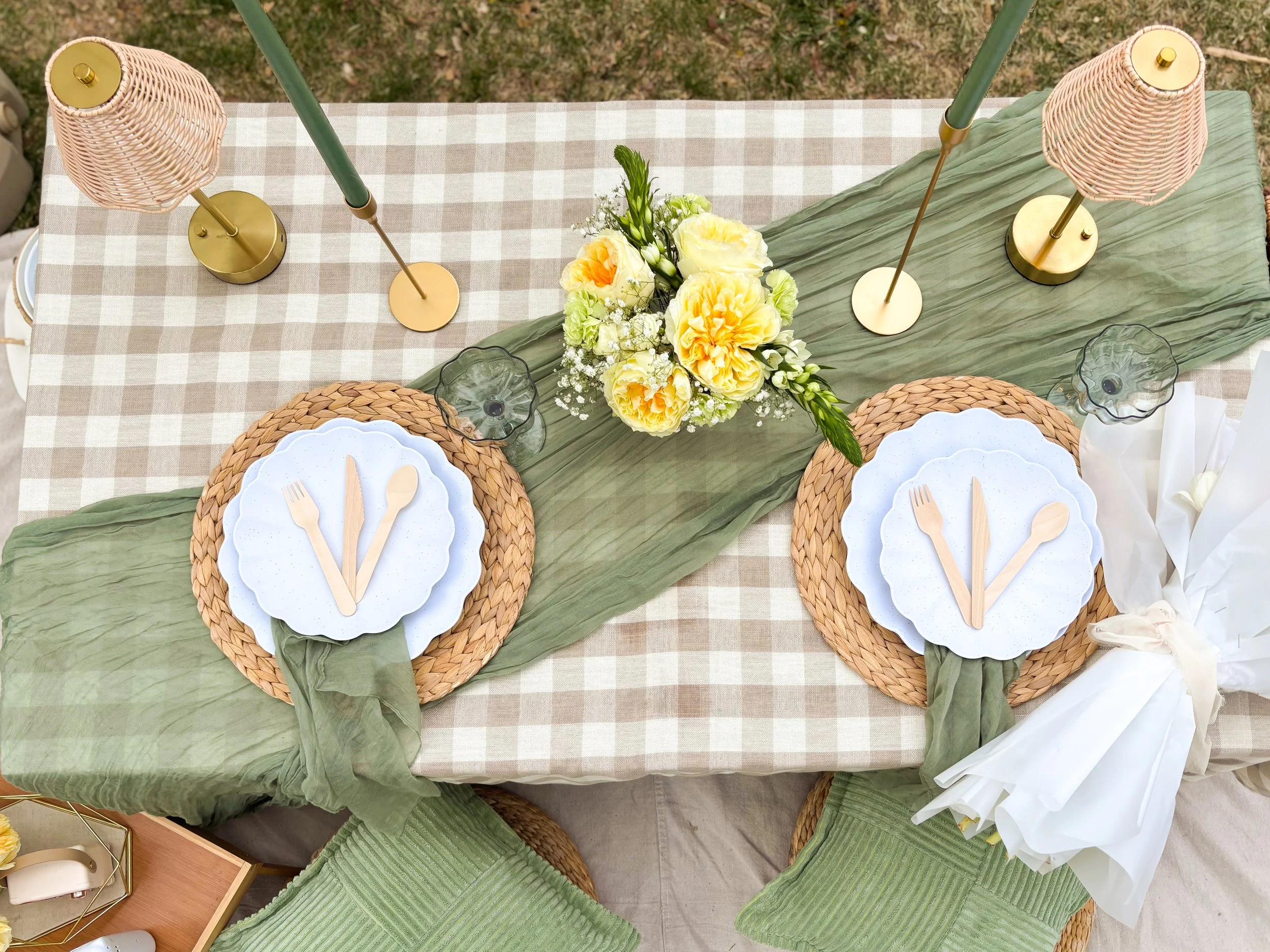 Overhead view of a table decorated with a green table runner, yellow floral centerpiece, wicker placemats, and white plates with wooden utensils. Wicker lamps and small vases are also on the table.