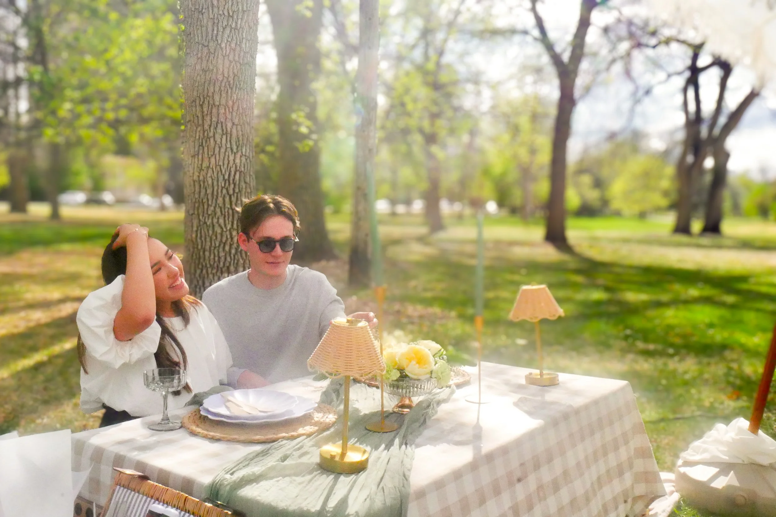 People enjoying a picnic in Denver City Park with trees and sunlight, sitting at a table with food, drinks, and decorative lamps.