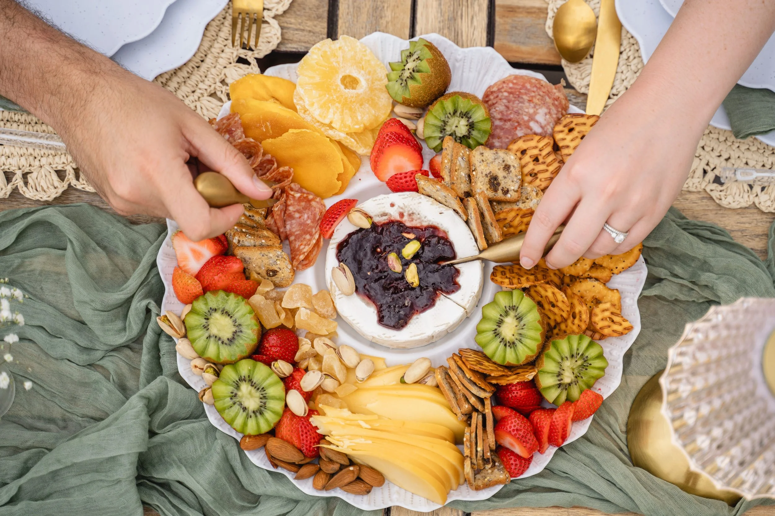 A round platter with assorted fruits, cheeses, crackers, and nuts, set on a green cloth with gold cutlery nearby.