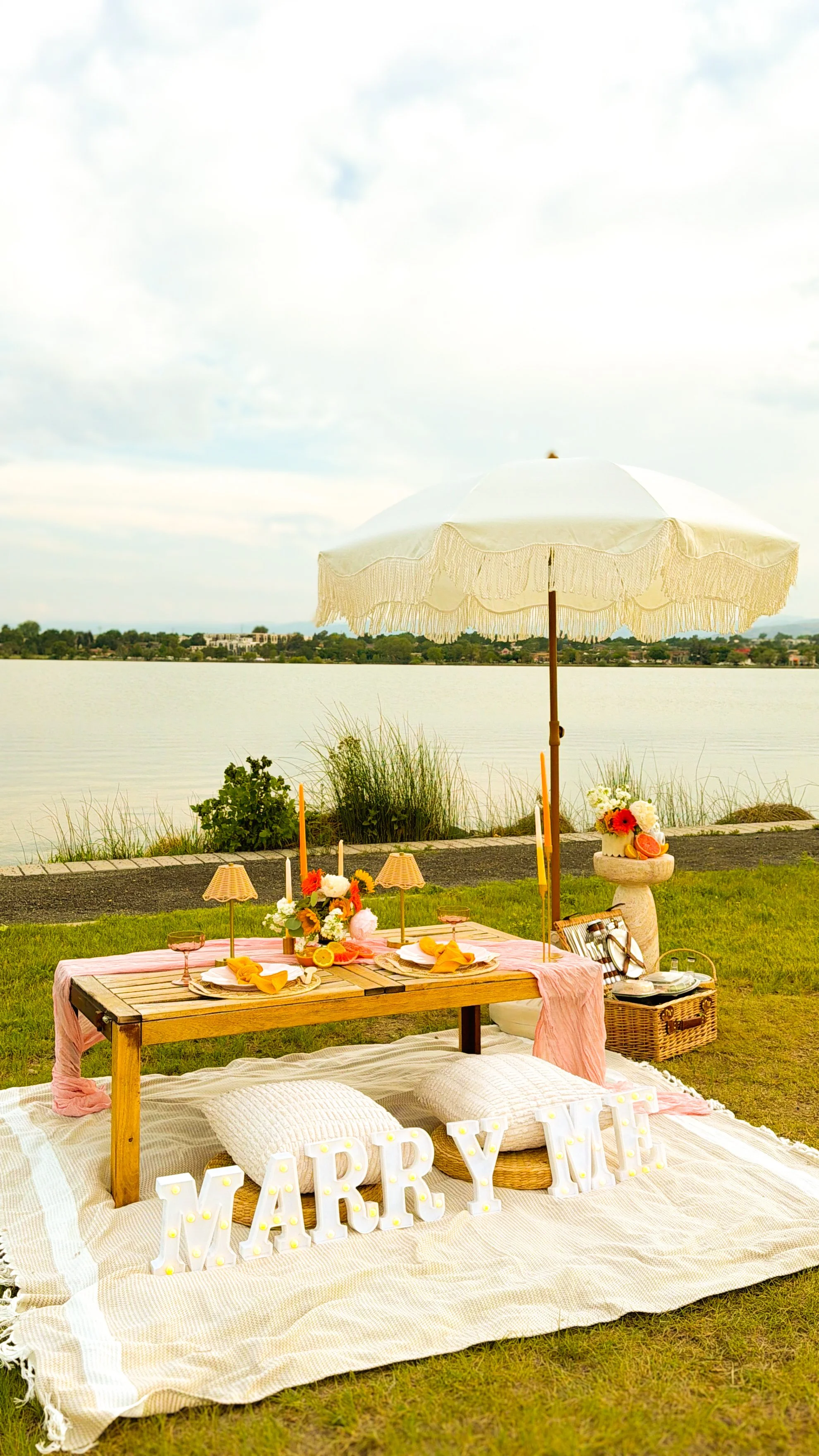 A picnic setup by a lake with a large white umbrella, a wooden table with pink tablecloth, flowers, candles, and plates, with large decorative letters spelling 'MARRY ME' on a white blanket in the foreground.