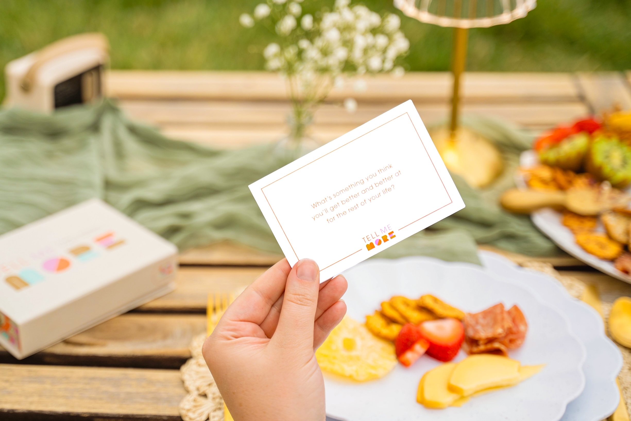 Person holding a white card at a picnic table with a spread of cheese and crackers, flowers, and snacks in the background.