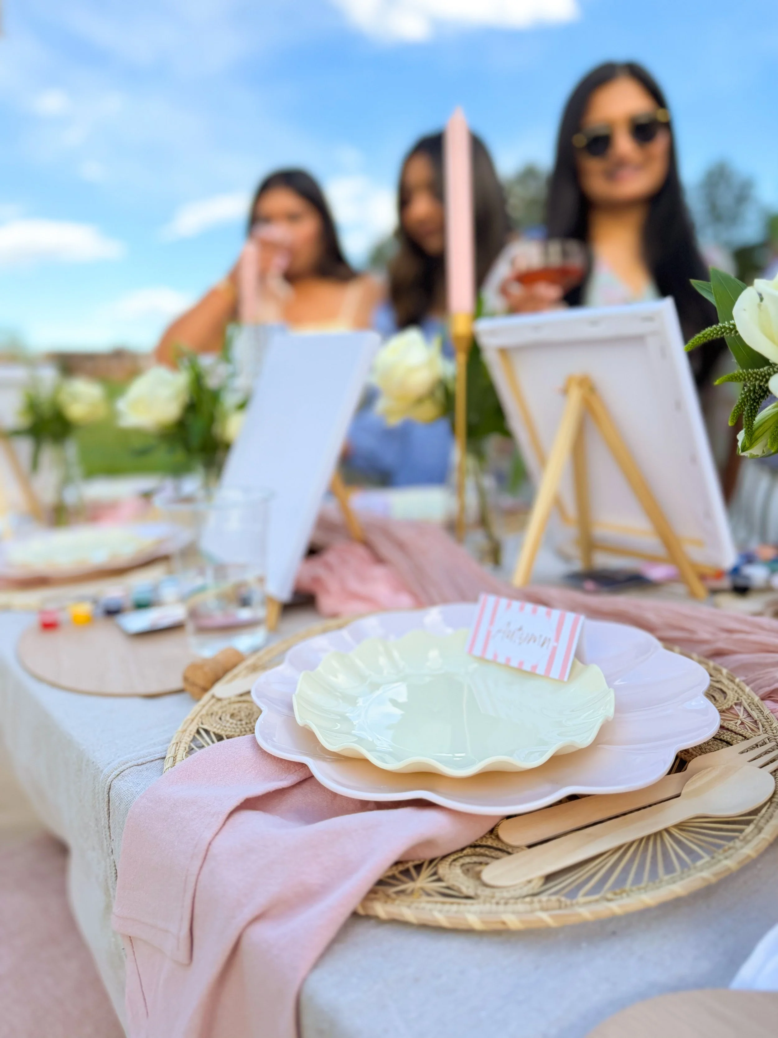 Close-up of a decorated table with a pink cloth, white plates, and wooden utensils, set outdoors with blurred women in the background