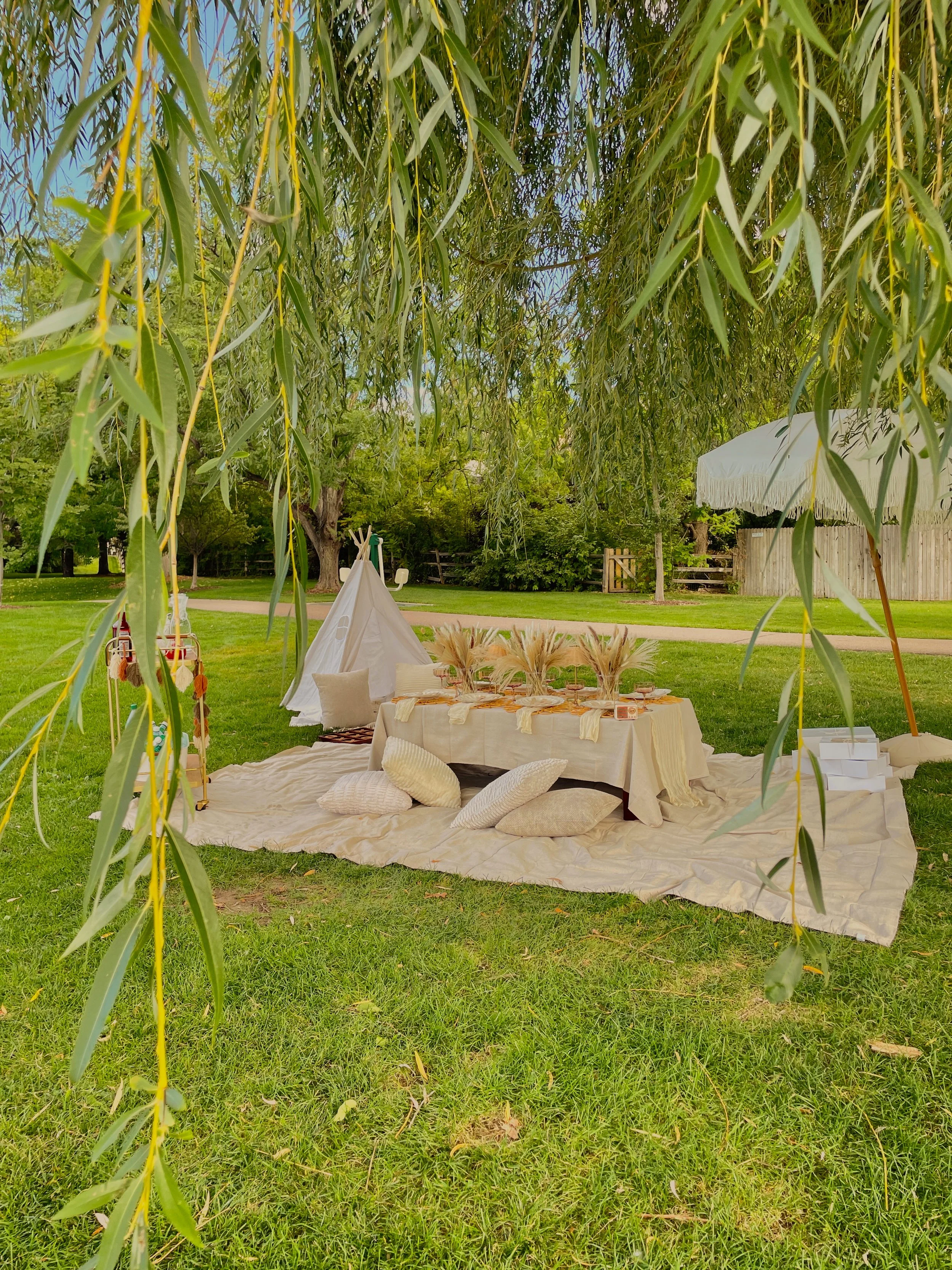 A picnic setup under a tree with a cream-colored blanket on the grass, surrounded by pillows, with a white outdoor umbrella and green trees in the background.