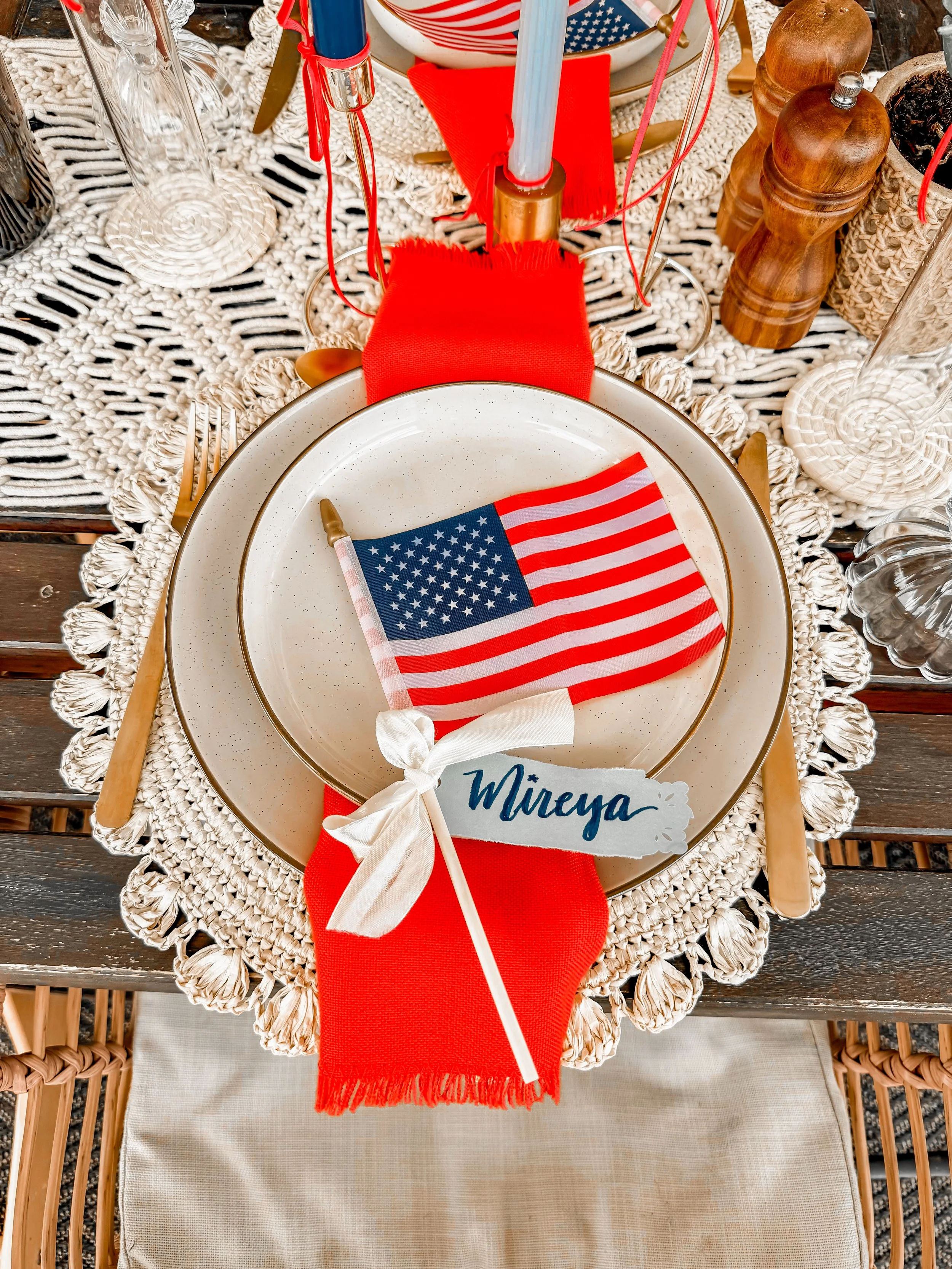 Table setting with a white plate, a small American flag, and a name tag with the name 'Mireya,' on a red napkin with a woven tablecloth, surrounded by glassware and utensils.