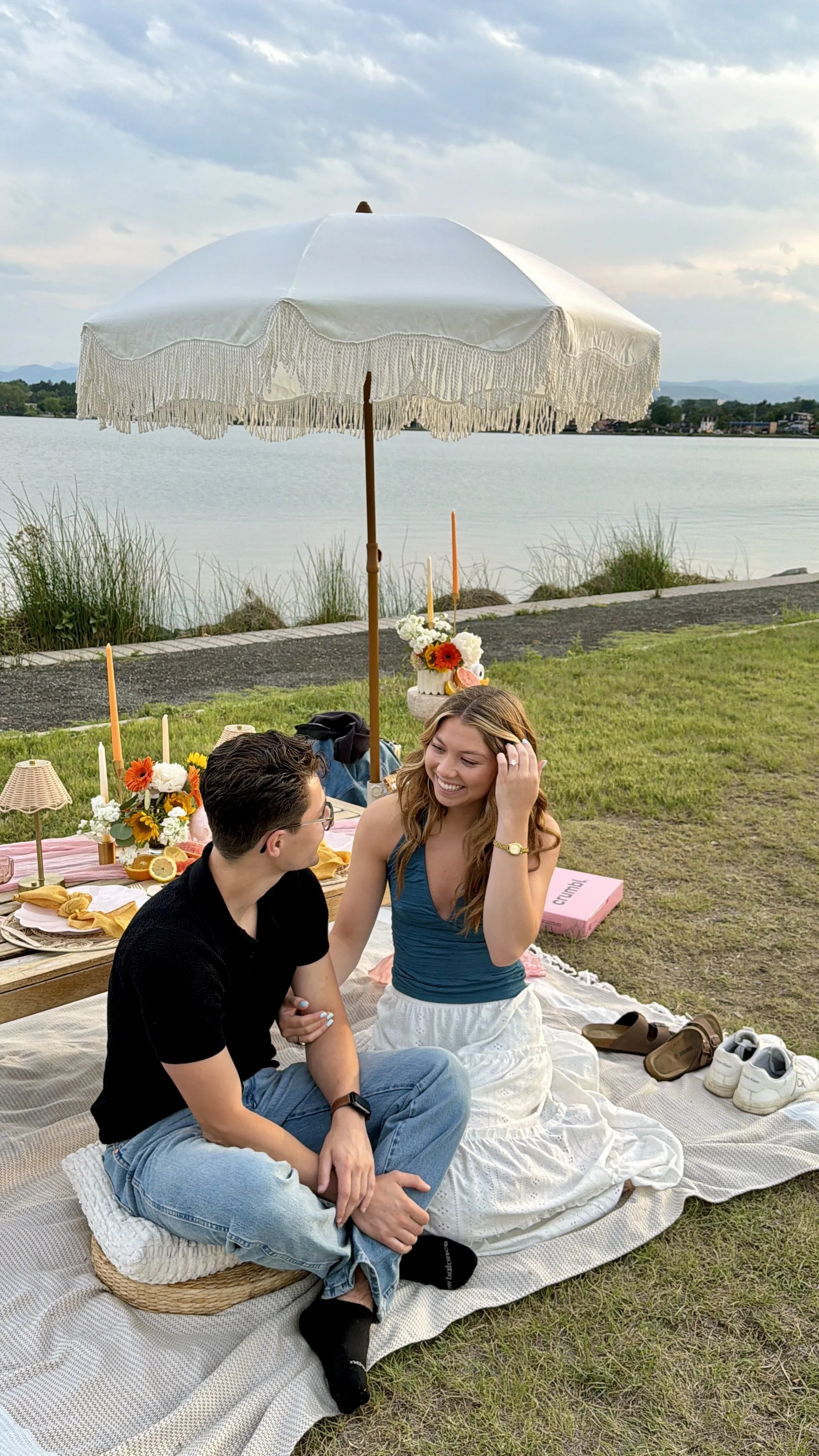 A couple sitting on a blanket by the water during a picnic, with flowers, candles, and an umbrella decorated for a proposal, smiling and engaging in conversation.