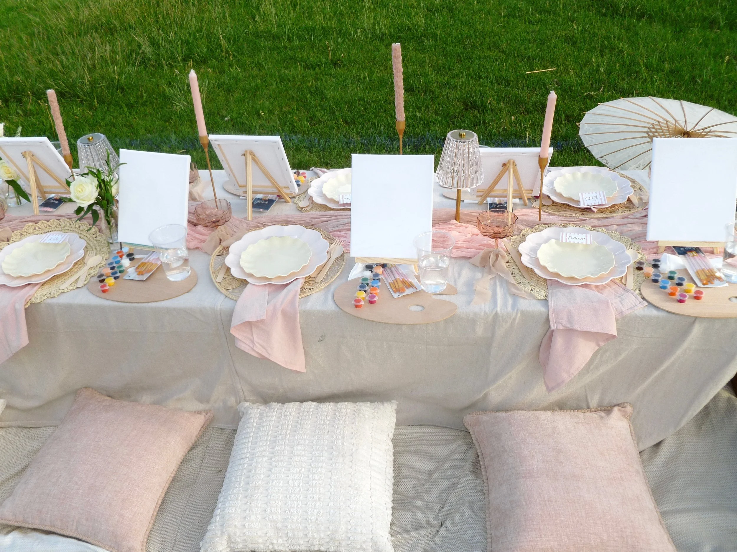 A table set for a celebration with pink candles, white flowers, and decorative plates, on a pink table runner, surrounded by pink and white cushions, with a grassy outdoor background.