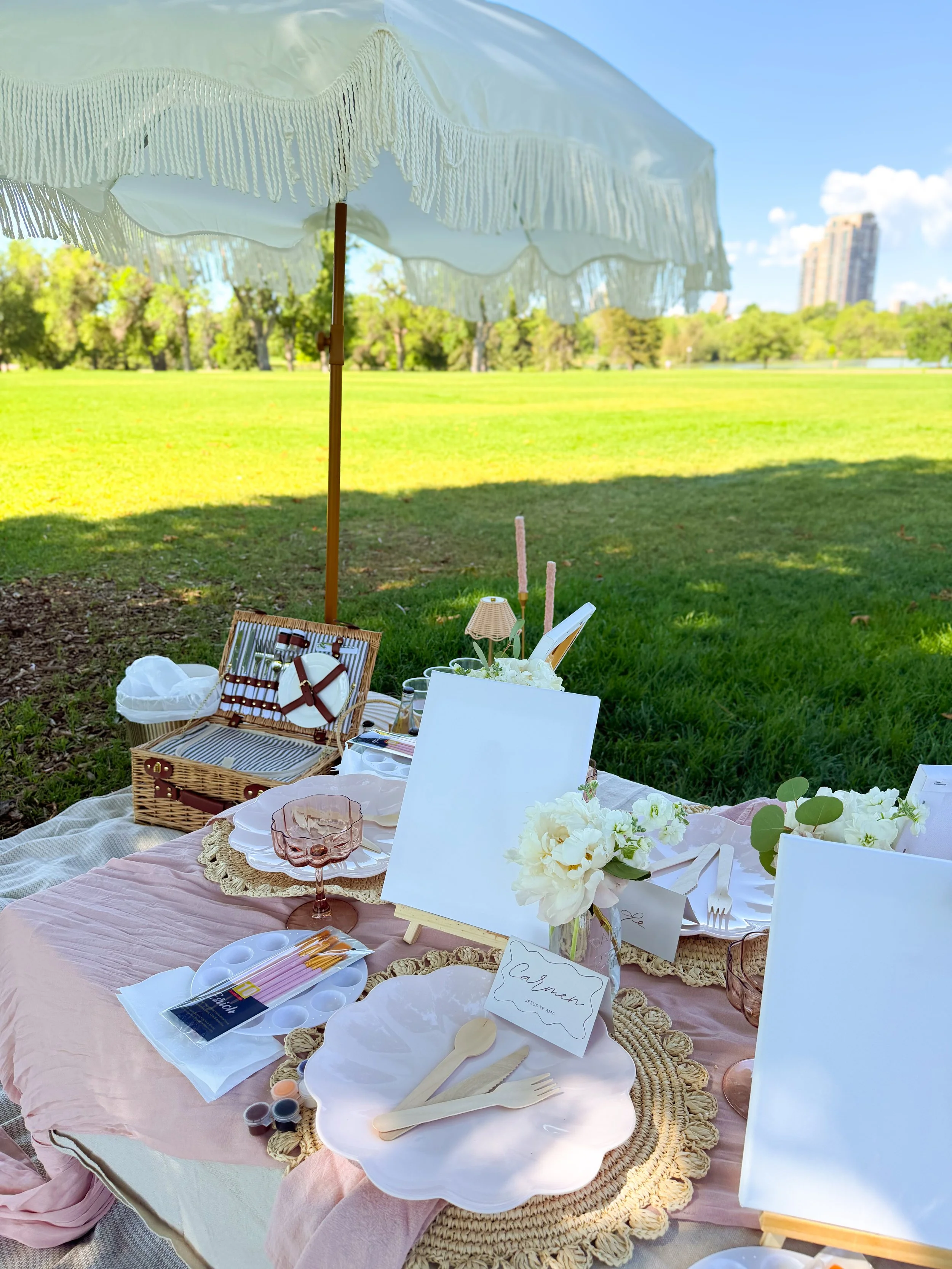 A bespoke picnic setup on a green lawn with a pink tablecloth, white floral centerpieces, and blank canvases under a white fringe umbrella with a city skyline in the background.