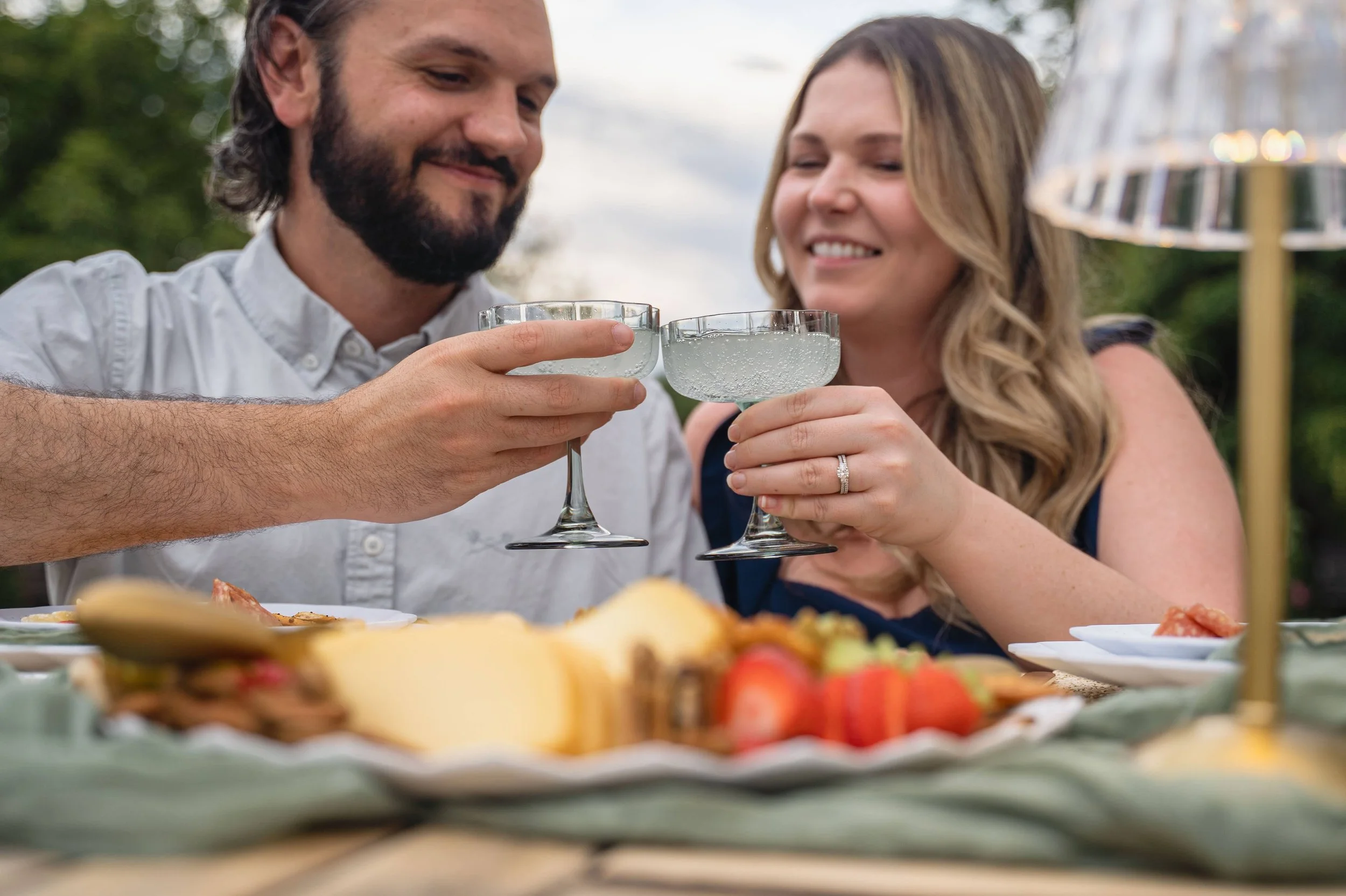 A man and woman smiling and toasting with drink glasses at an outdoor gathering, with plates of food on the table.
