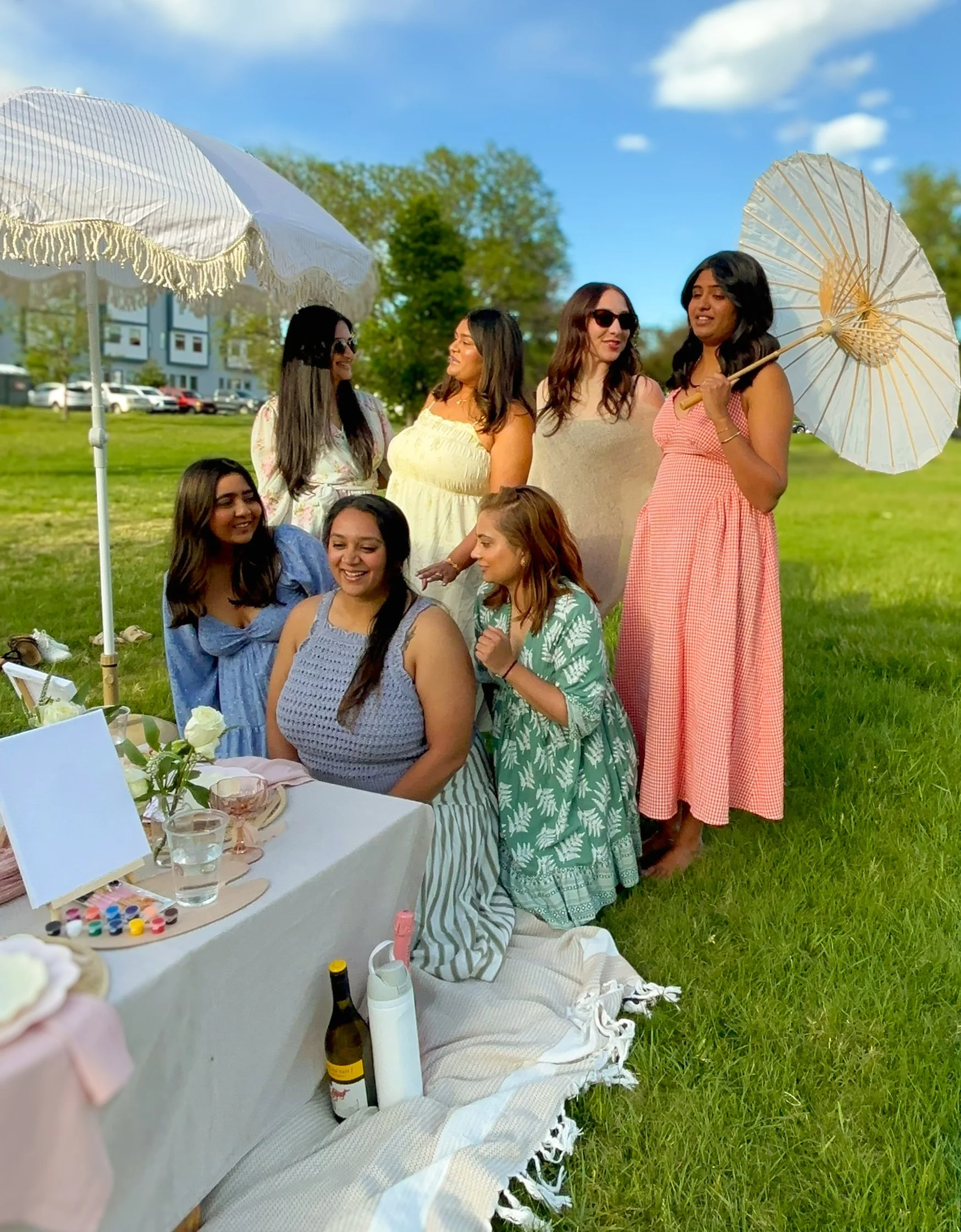 Group of women in dresses and sunglasses standing and sitting outdoors on grass, sitting at a luxury picnic table, with a large white parasol and smaller parasol in the background under a blue sky.