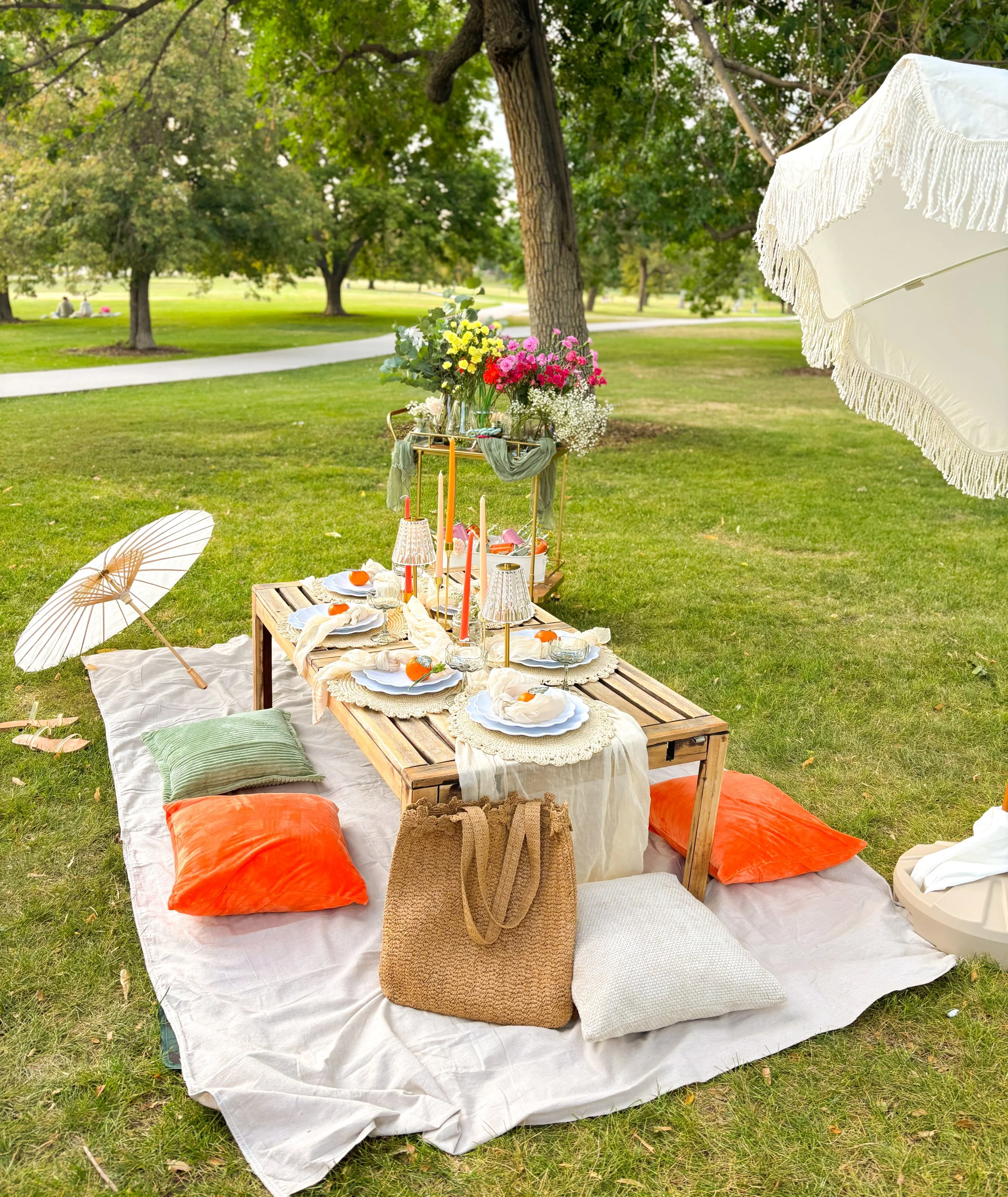 A picnic setup on the grass with a low wooden table, pillows, blankets, candles, flowers, and umbrellas in a park with trees and a walking path in the background.