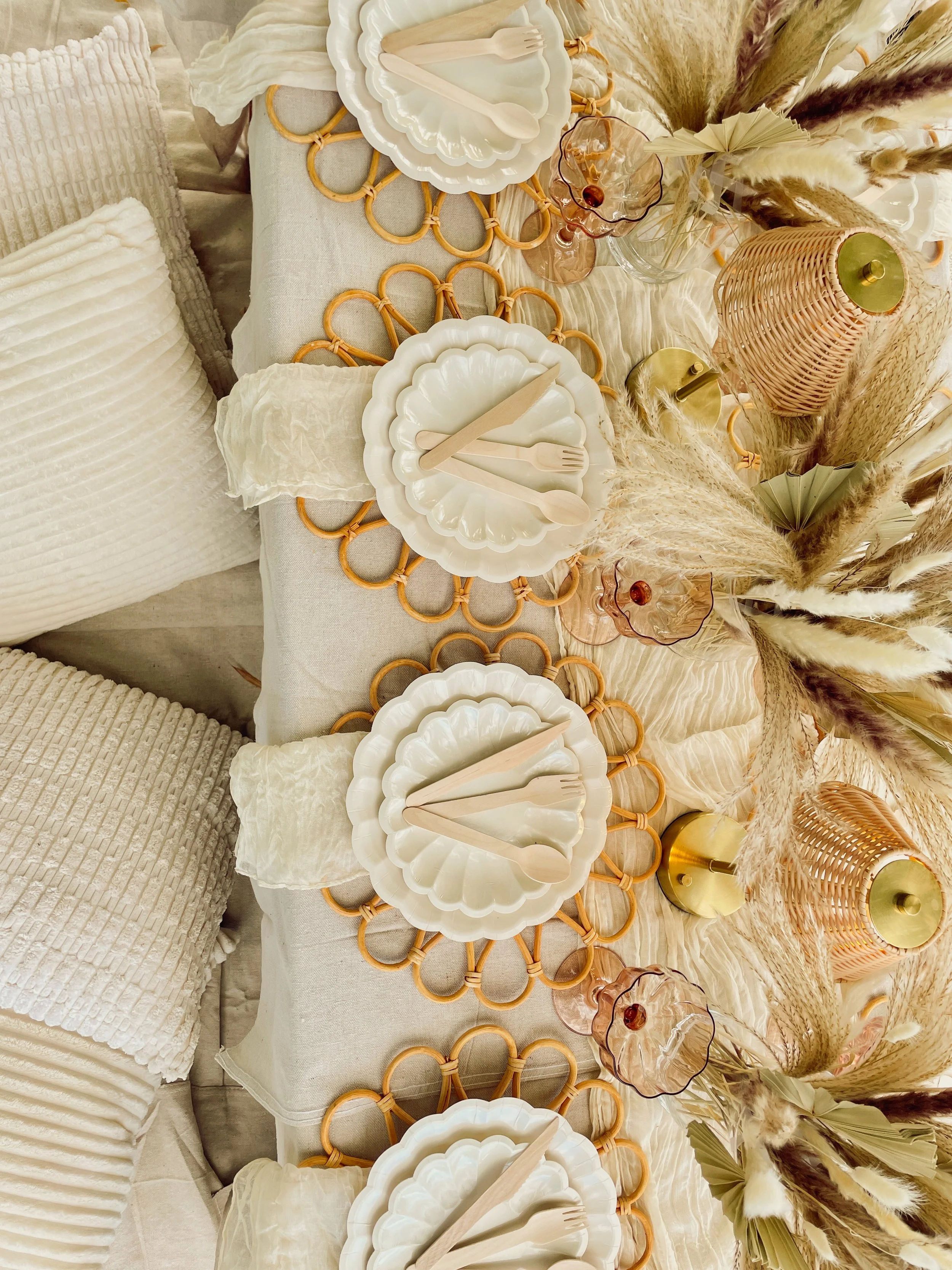 Decorative dining table setting with white scalloped plates, wooden utensils, pink and clear glassware, woven rattan placemats, and dried pampas grass floral arrangement in neutral tones.