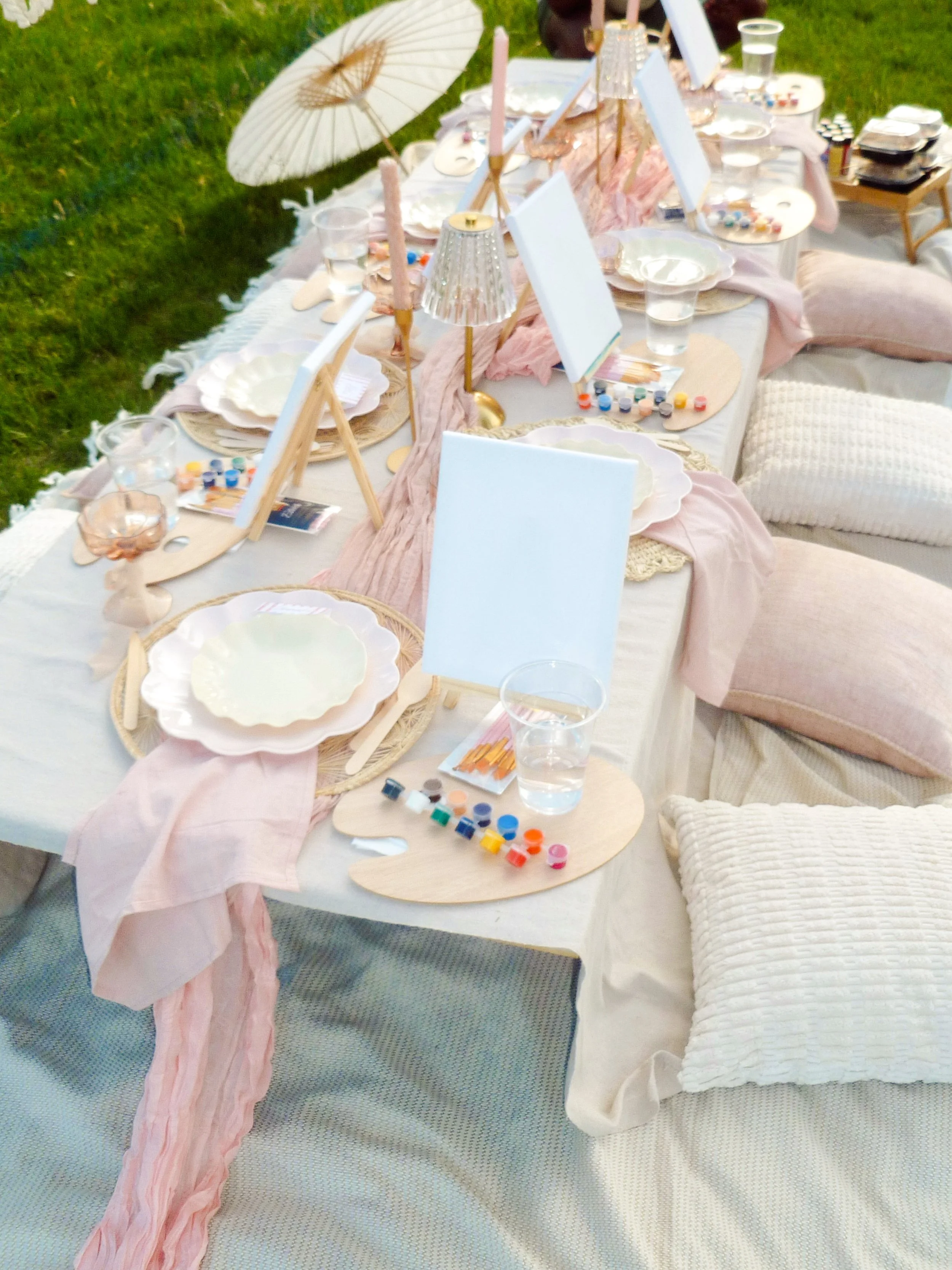 Decorative outdoor table setup with pink and white accents, including plates, glasses, candles, small easels with blank white boards, and art supplies like paint and brushes, surrounded by cushions and a pink fabric runner, on a grassy area.