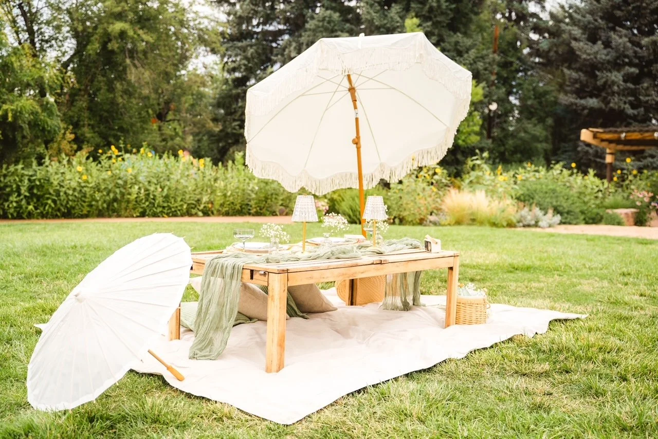 Picnic setup on a grassy lawn with a low wooden table, two white umbrellas, and a white cloth underneath. The table is decorated with small lamps, glasses, and a green table runner, surrounded by pillows and a basket.