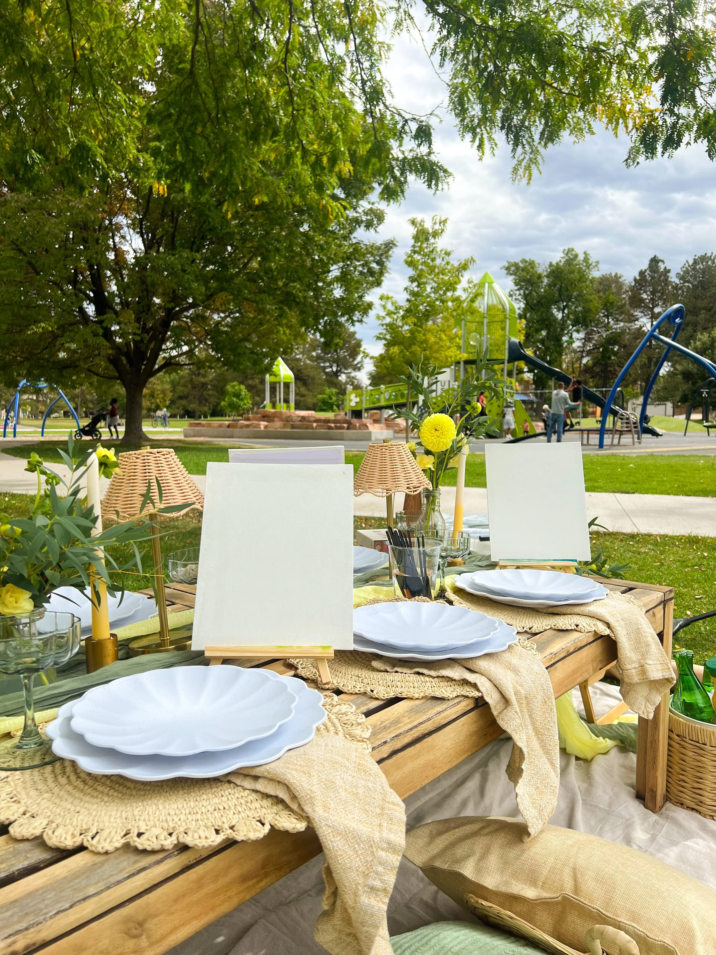 A picnic table set with white plates, glasses, and decorated with yellow flowers, situated outdoors in a park with trees, playground equipment, and people in the background.