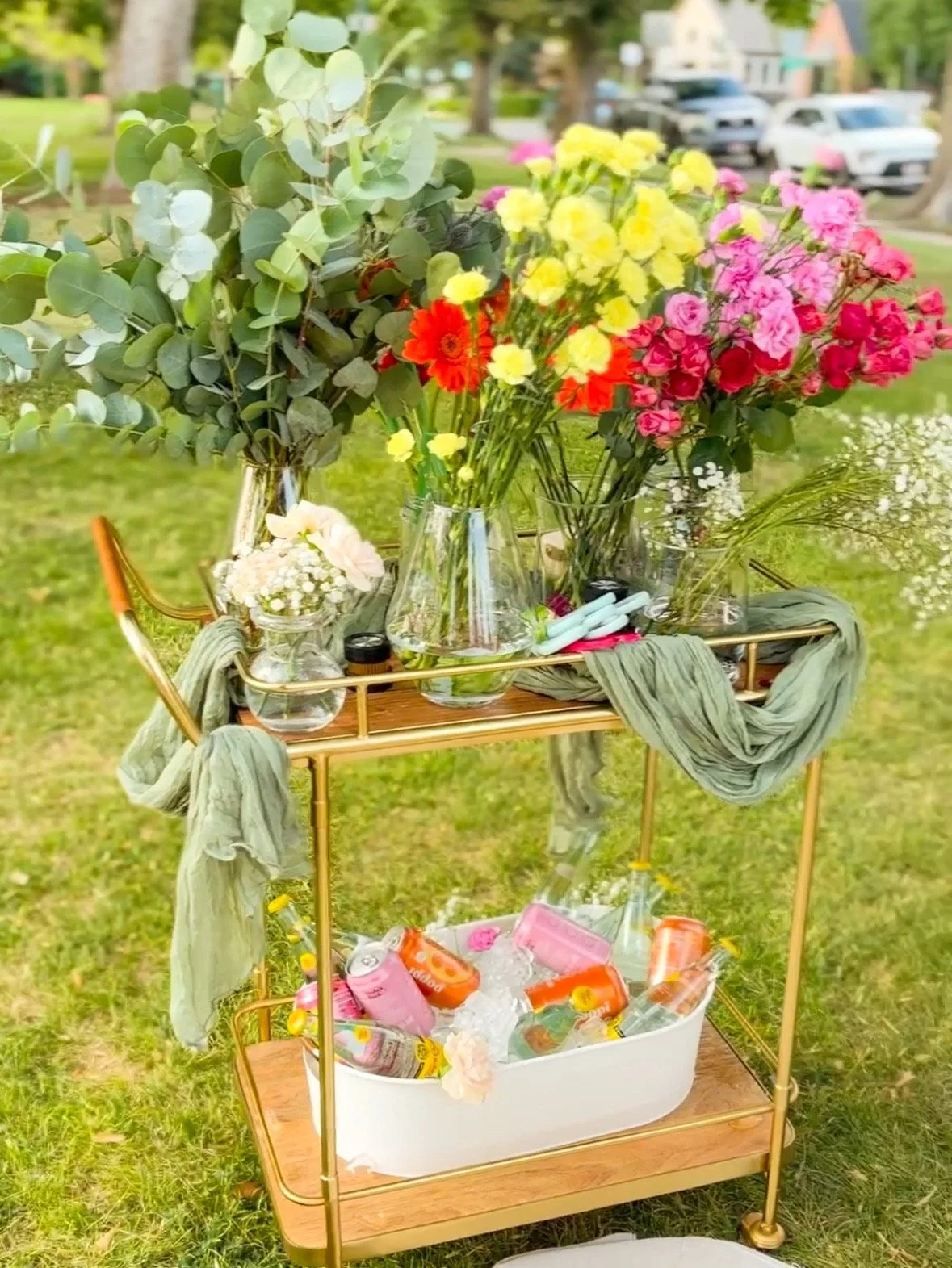 A wooden bar cart with various floral arrangements in glass vases and bottles, and drinks in cans and bottles, set outdoors on grass.