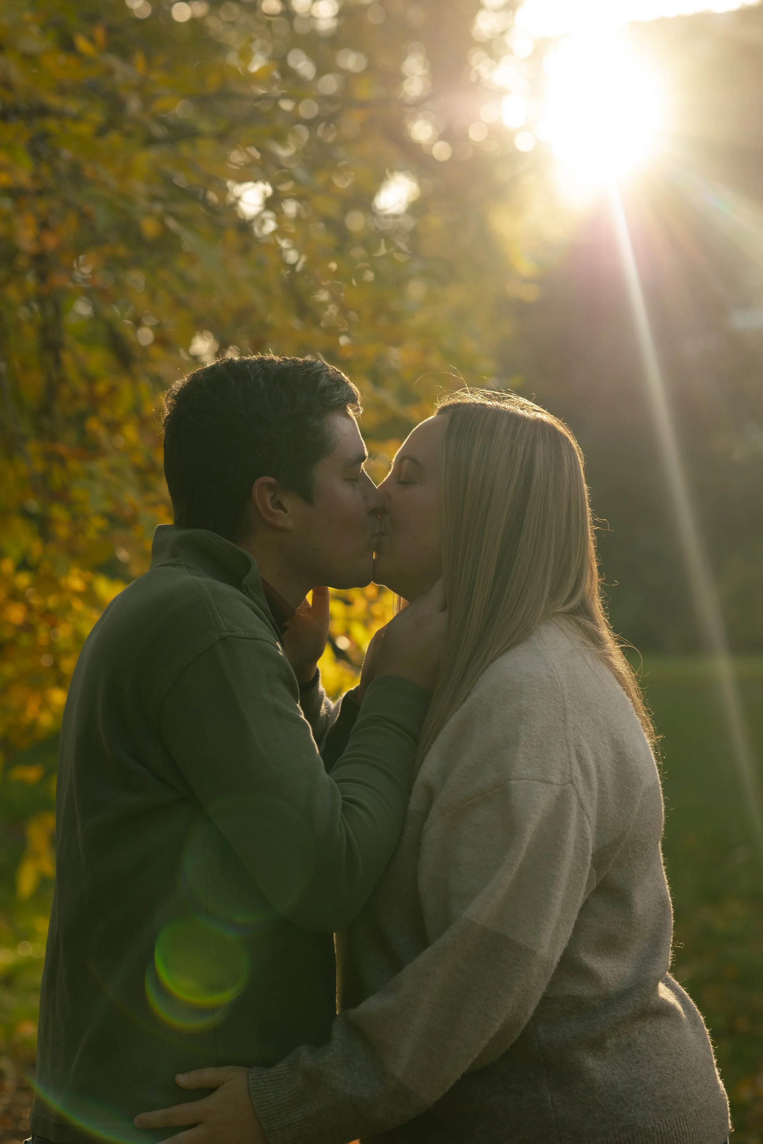A couple kisses outdoors with sunlight shining through trees in the background.