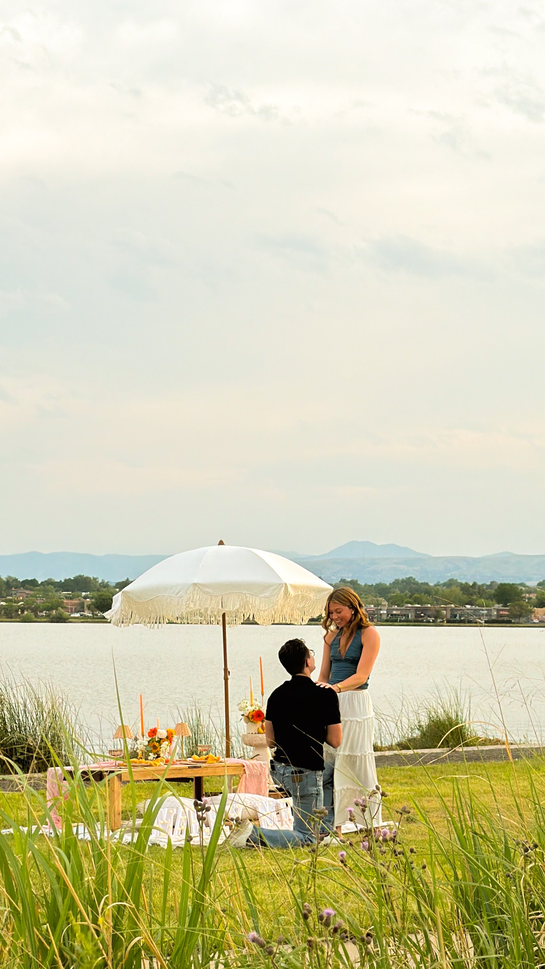 A couple is having a romantic moment outdoors by a lake with a scenic mountain view in the background. The woman is standing and smiling at the man who is kneeling in front of her, holding her hands. An umbrella provides shade, and a picnic setup wit
