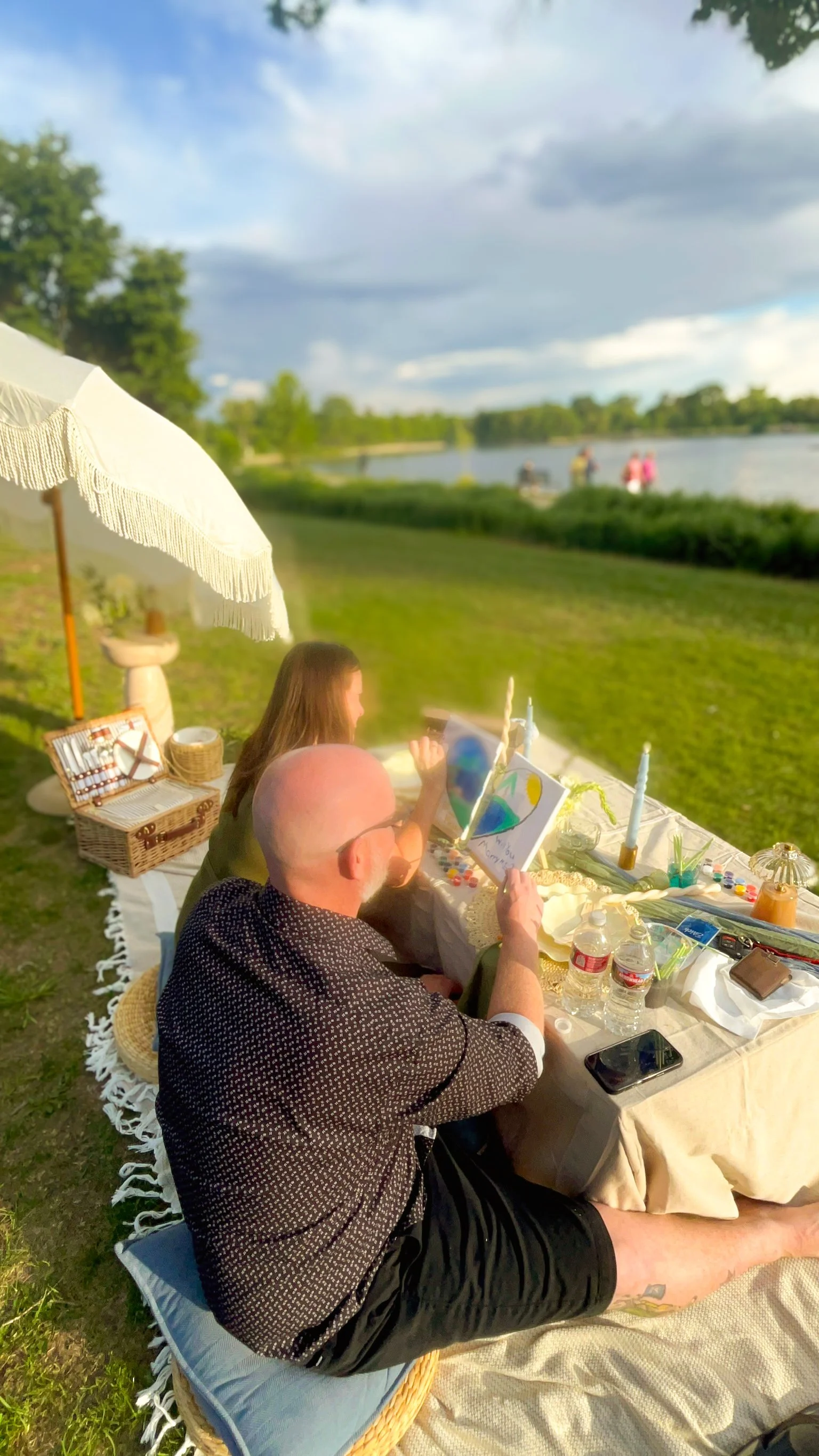 Two people sit at a picnic table with trees and a cloudy sky in the background. One person is painting on a small canvas, and the other person is looking at the painting. The table is decorated with various art supplies, candles, and water bottles. A