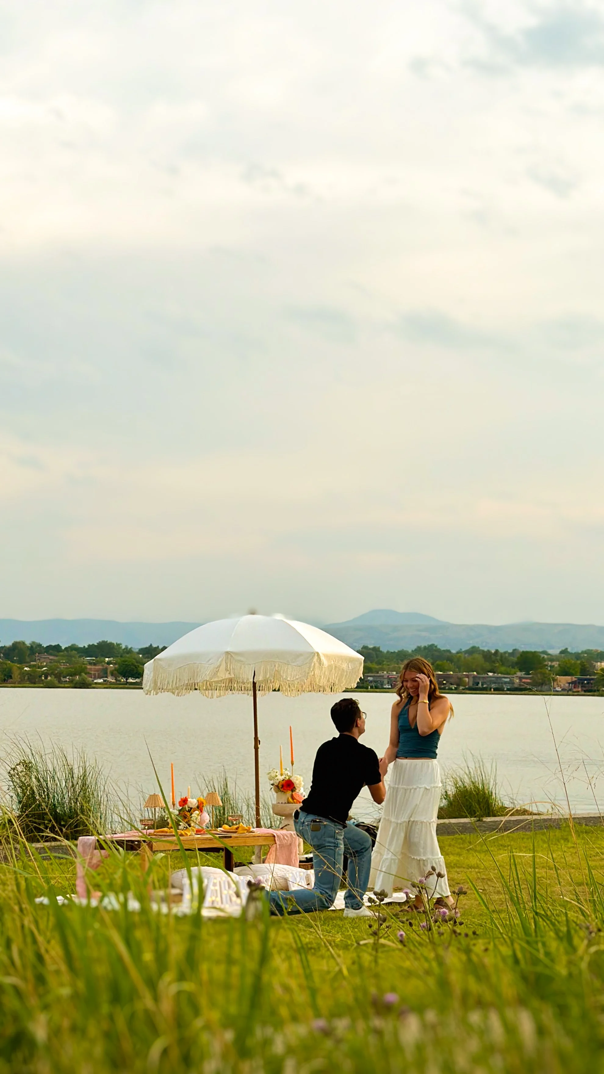 A couple has a proposal by the lake during a picnic, with a man kneeling and proposing to a woman, surrounded by flowers, a blanket, and an umbrella on a grassy area with mountains and water in the background.