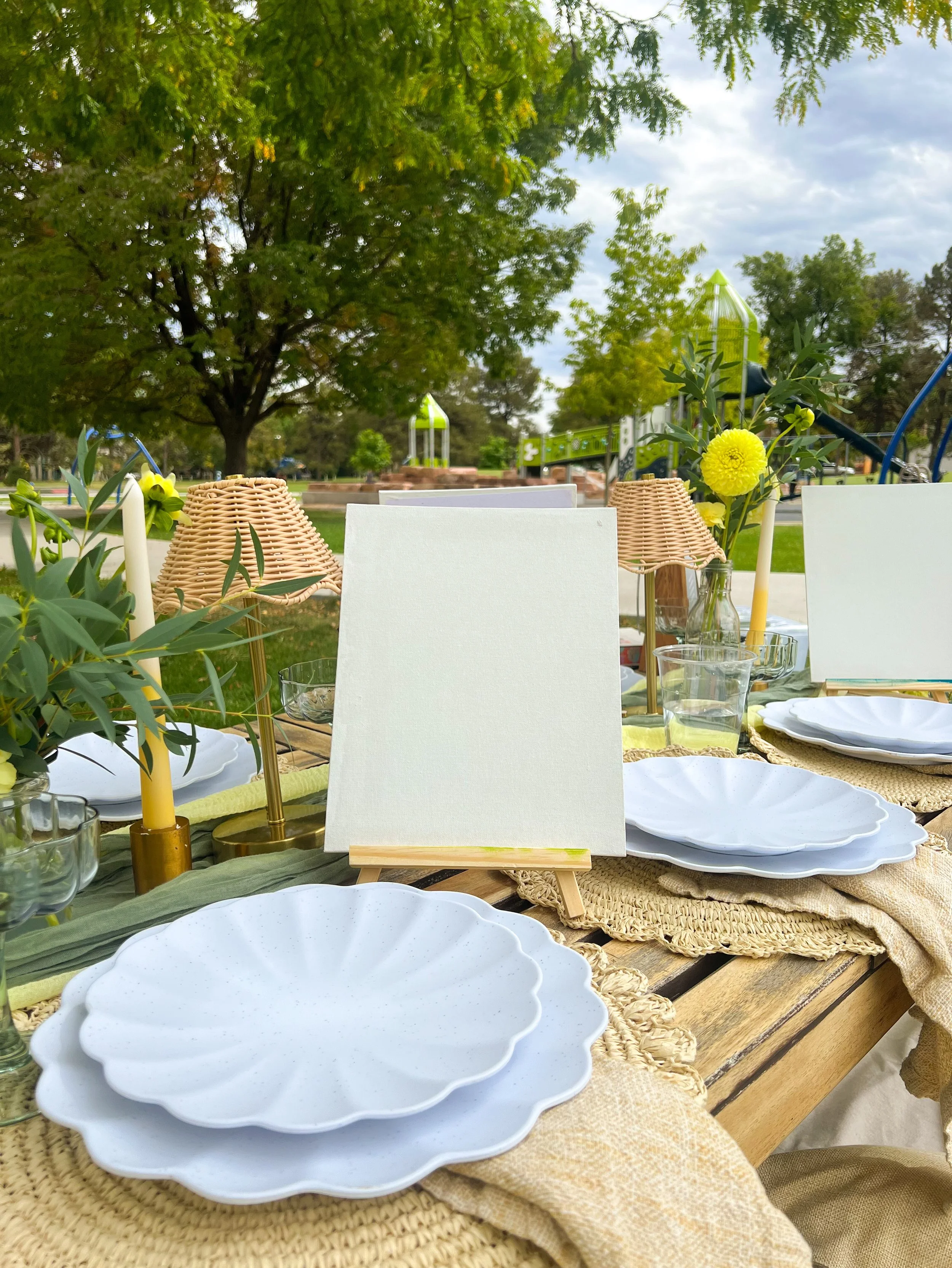 Outdoor table decorated for a gathering with white plates, yellow flowers, candles, glasses, and a canvas on a stand, set in a park with large trees and a playground in the background.