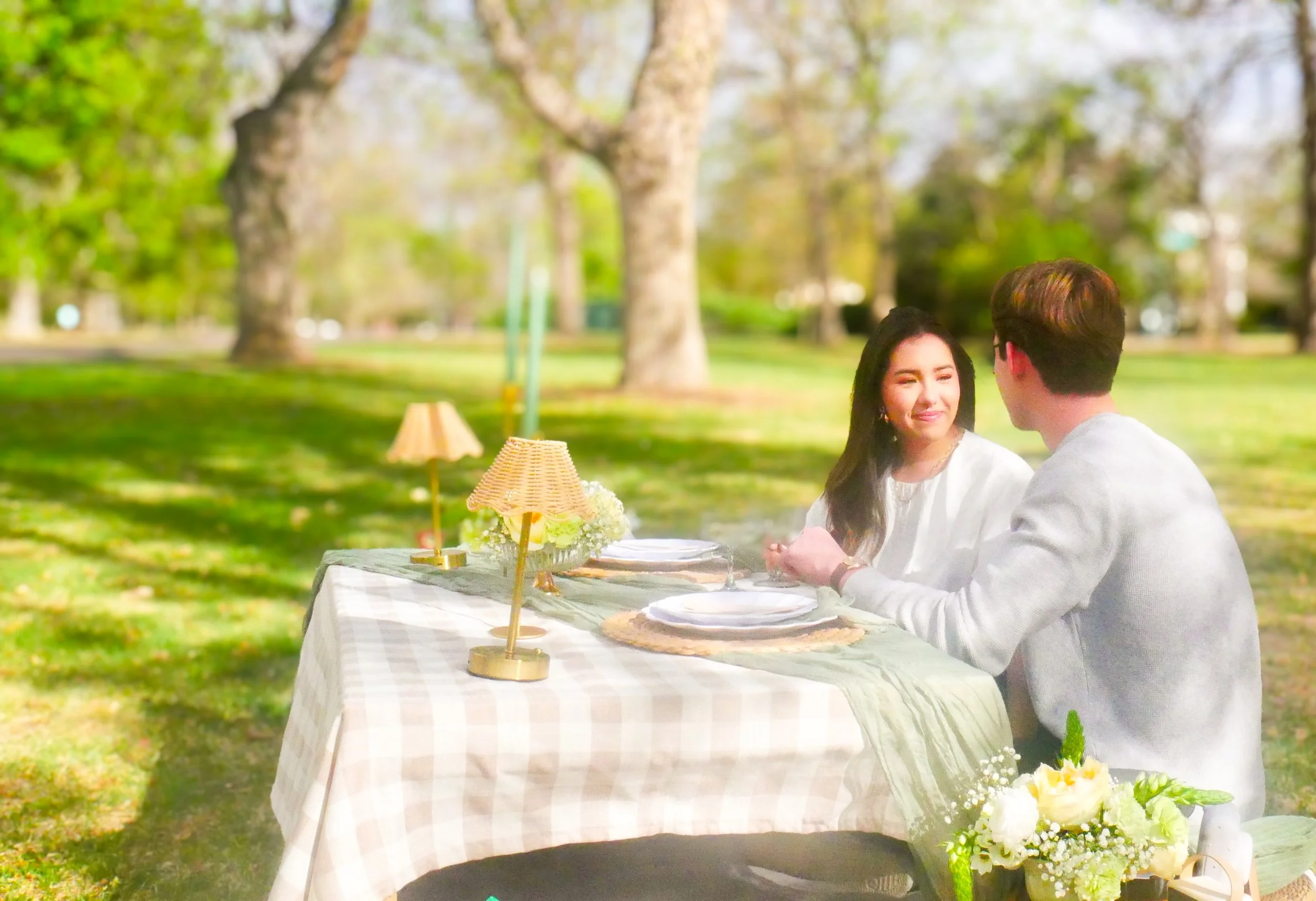 A couple sitting at a table outdoors in a park, engaged in a conversation. The table is covered with a checkered tablecloth and decorated with flowers and lamps.