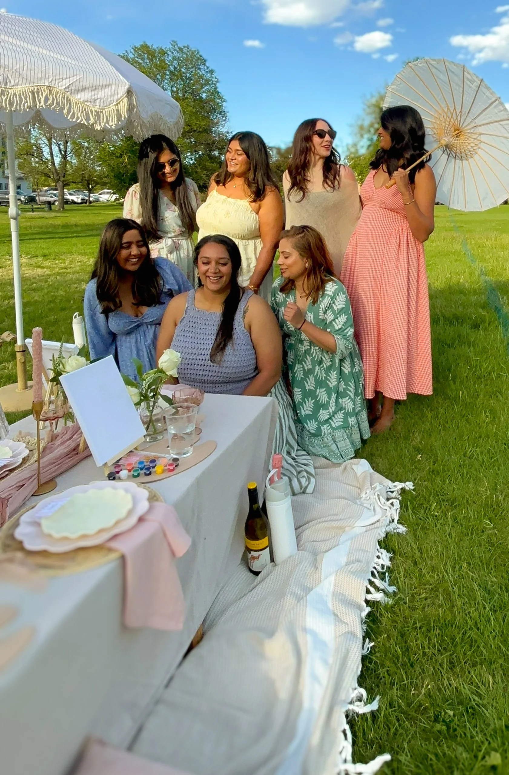 Group of seven women gathered around a decorated table outdoors during the day, with trees and a grassy field in the background. Some women are standing, and others are sitting on the grass, smiling and enjoying a luxury picnic in Wash Park, Denver.