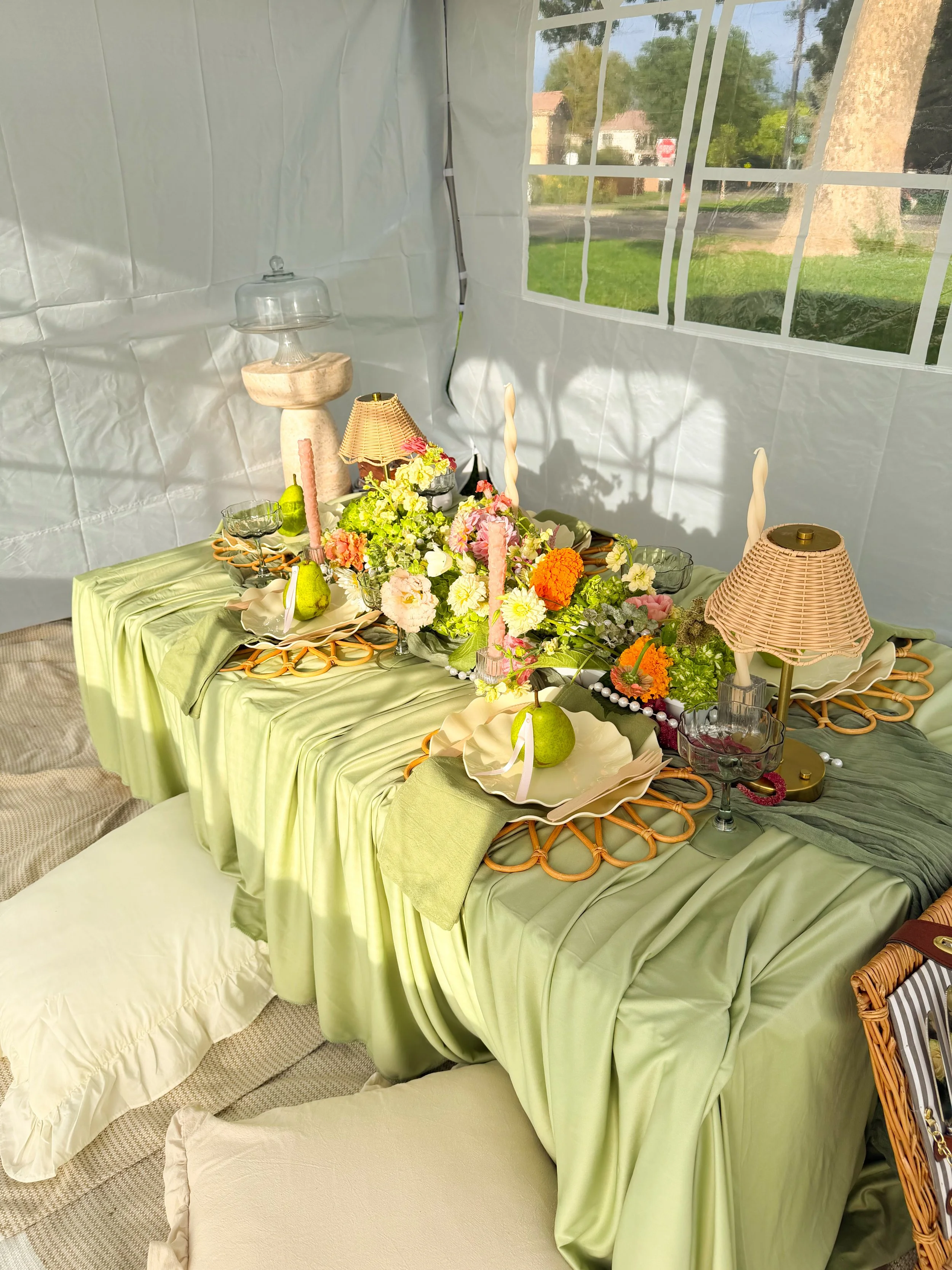 A decorated luxury picnic table with green and beige fabric tablecloths, floral arrangements, wicker and glass lamps, and plate settings.