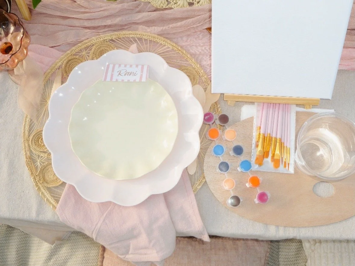 A luxury picnic table set up for a painting project with paint bottles, paintbrushes, a blank canvas, and decorative tray and tablecloths.