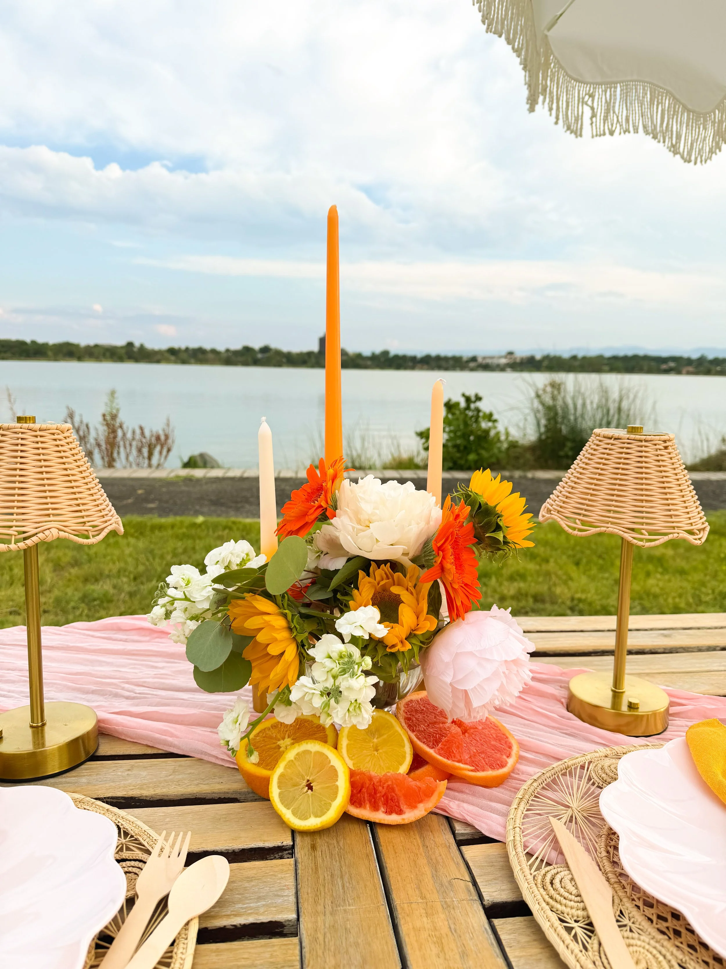 A table decorated with a pink tablecloth, a floral centerpiece, and sliced citrus fruits, set outdoors with a lake and cloudy sky in the background. There are wicker lamps and plates with bamboo utensils on the table.