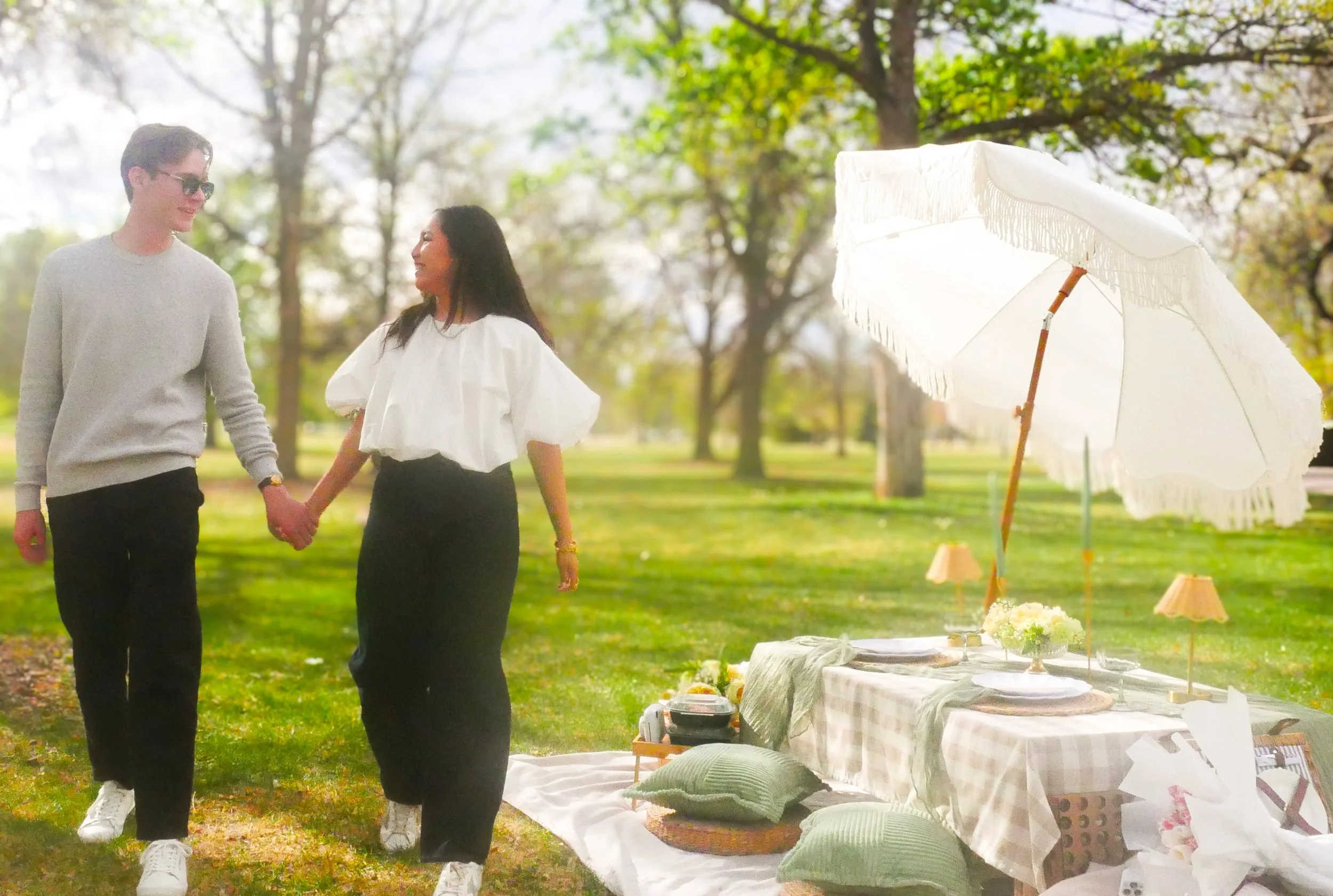 A couple holding hands and smiling outdoors near a table set for a celebration, with trees and sunlight in the background.