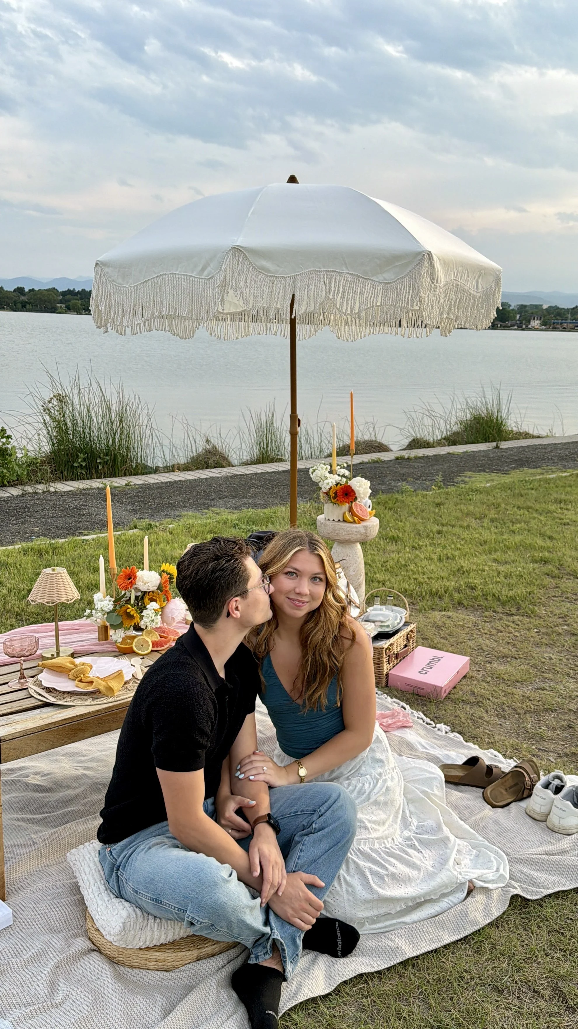 A couple sitting on a blanket during a picnic by a lake, with a white umbrella, flowers, fruits around them.