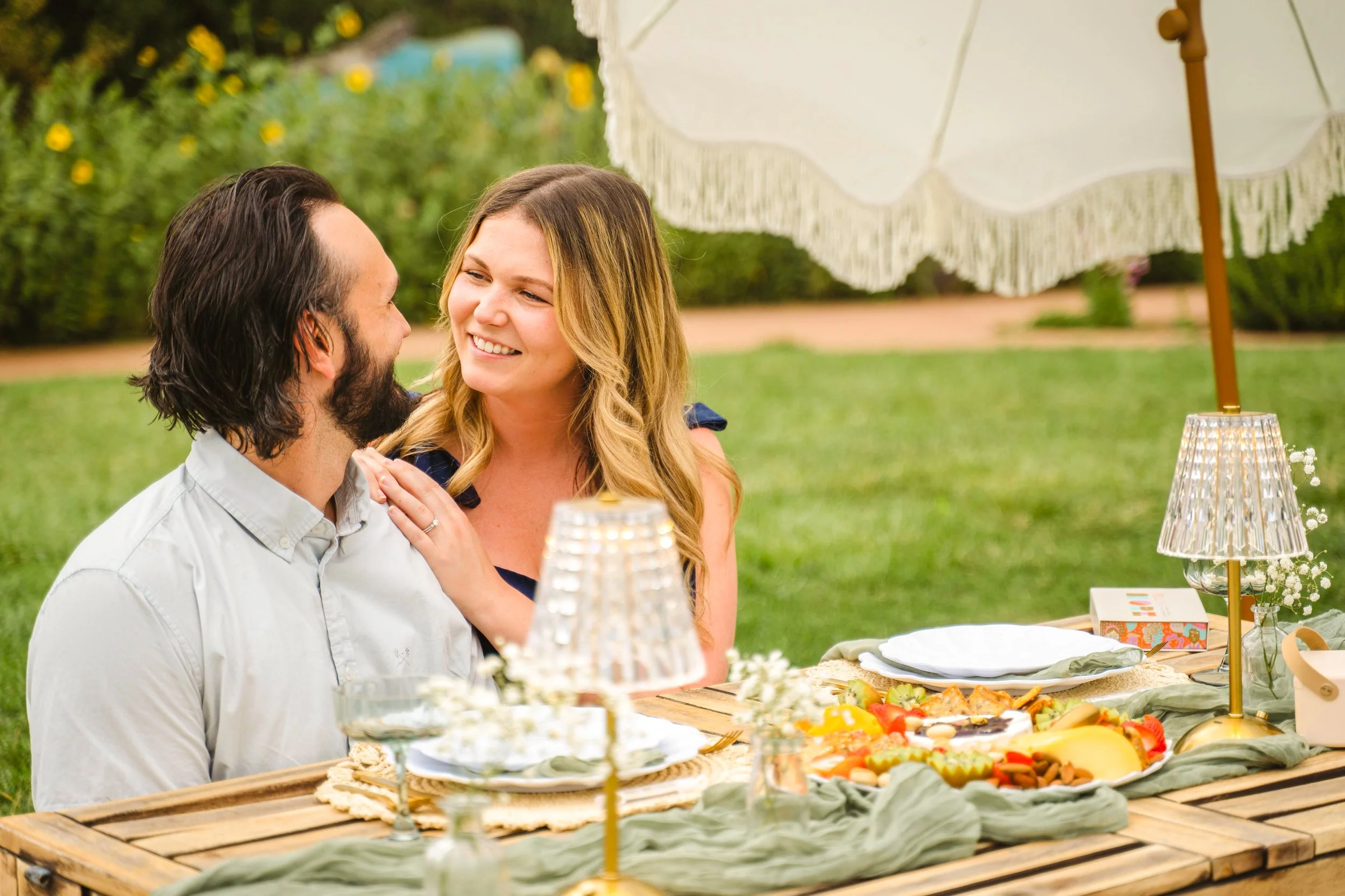 A man and woman smiling and looking at each other at an outdoor gathering with a table of food, including fruit and desserts, under a large white umbrella.