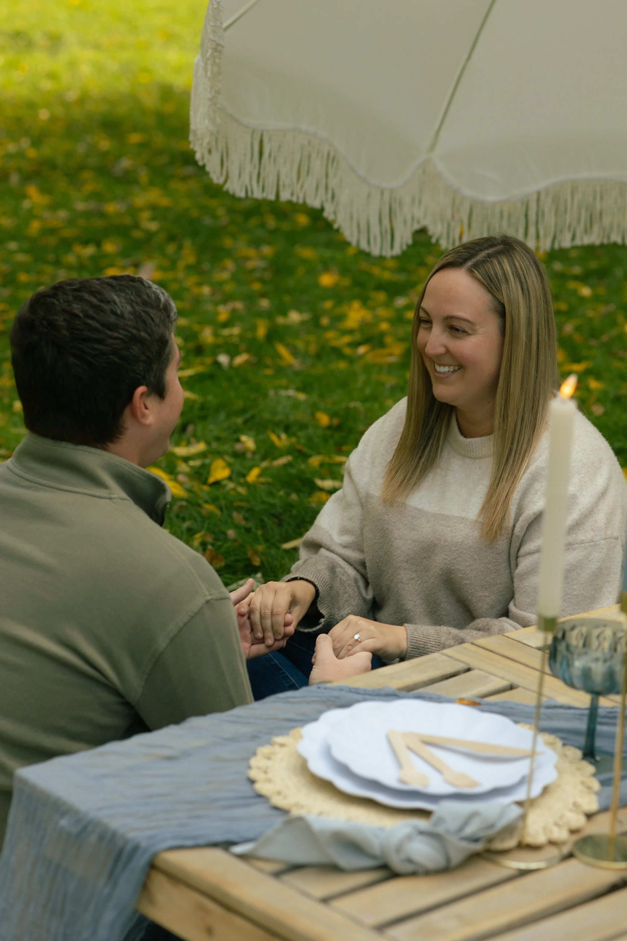 A woman and a man sitting at a table outdoors under a large beige umbrella, holding hands and smiling at each other, with a table of food in the foreground.