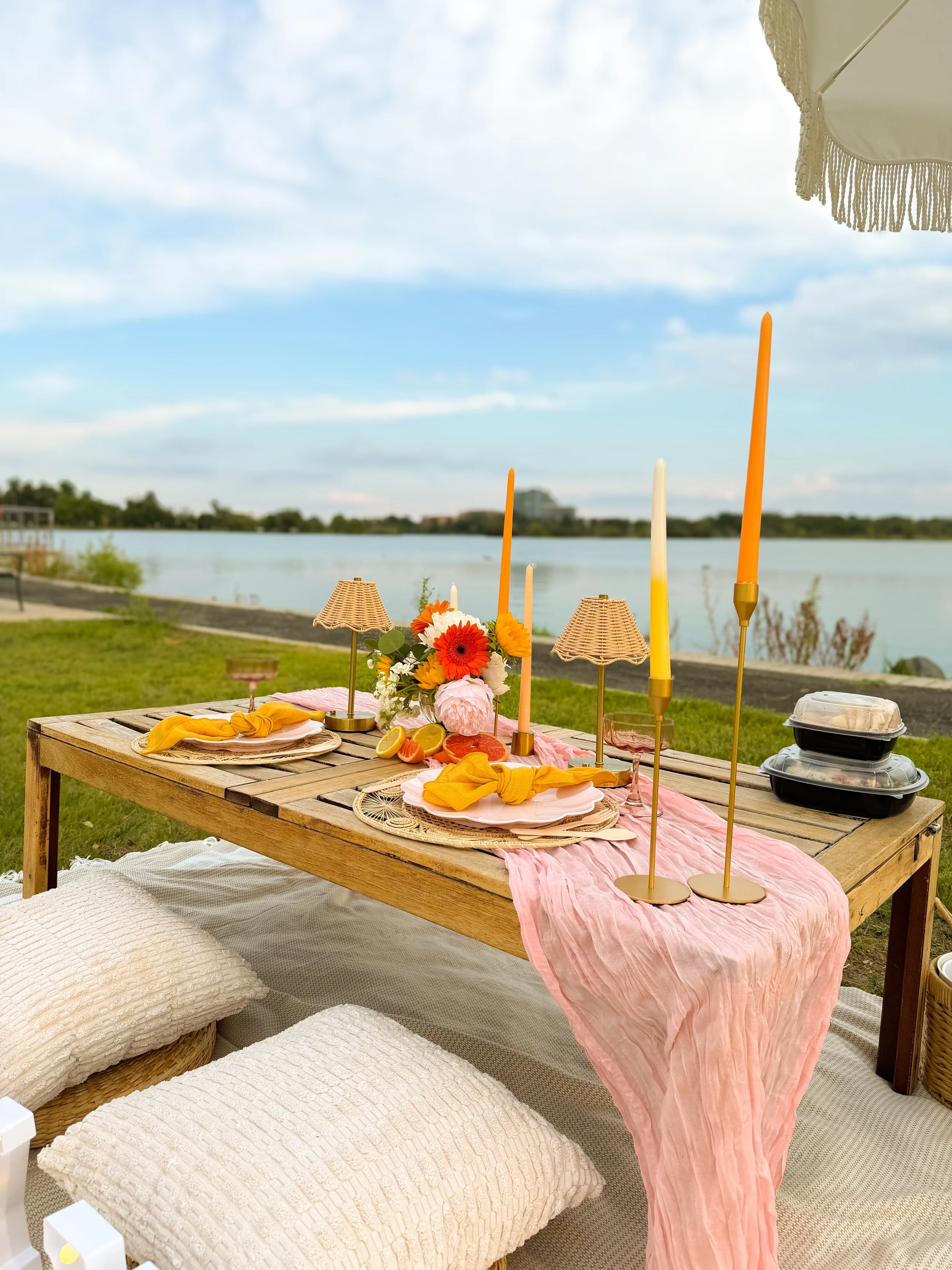 A lakeside outdoor table set for a gathering with pink tablecloth, floral centerpiece, candles, plates, and yellow napkins, with cushions on the ground and a lake with trees and buildings in the background.