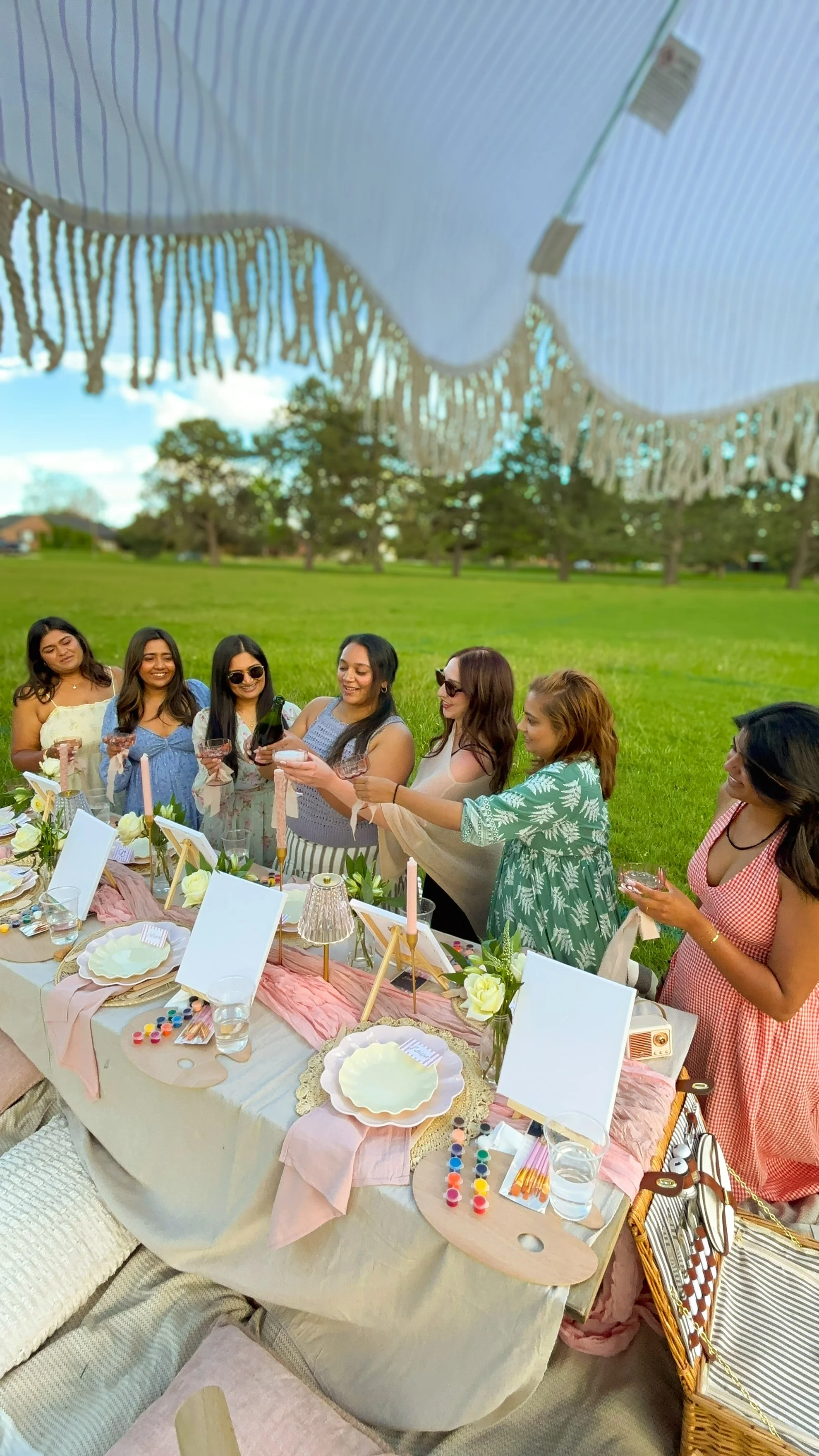 Group of women celebrating a bachelorette at a luxury picnic table decorated with pink fabric, flowers, candles, and tableware, with a grassy field and trees in the background.