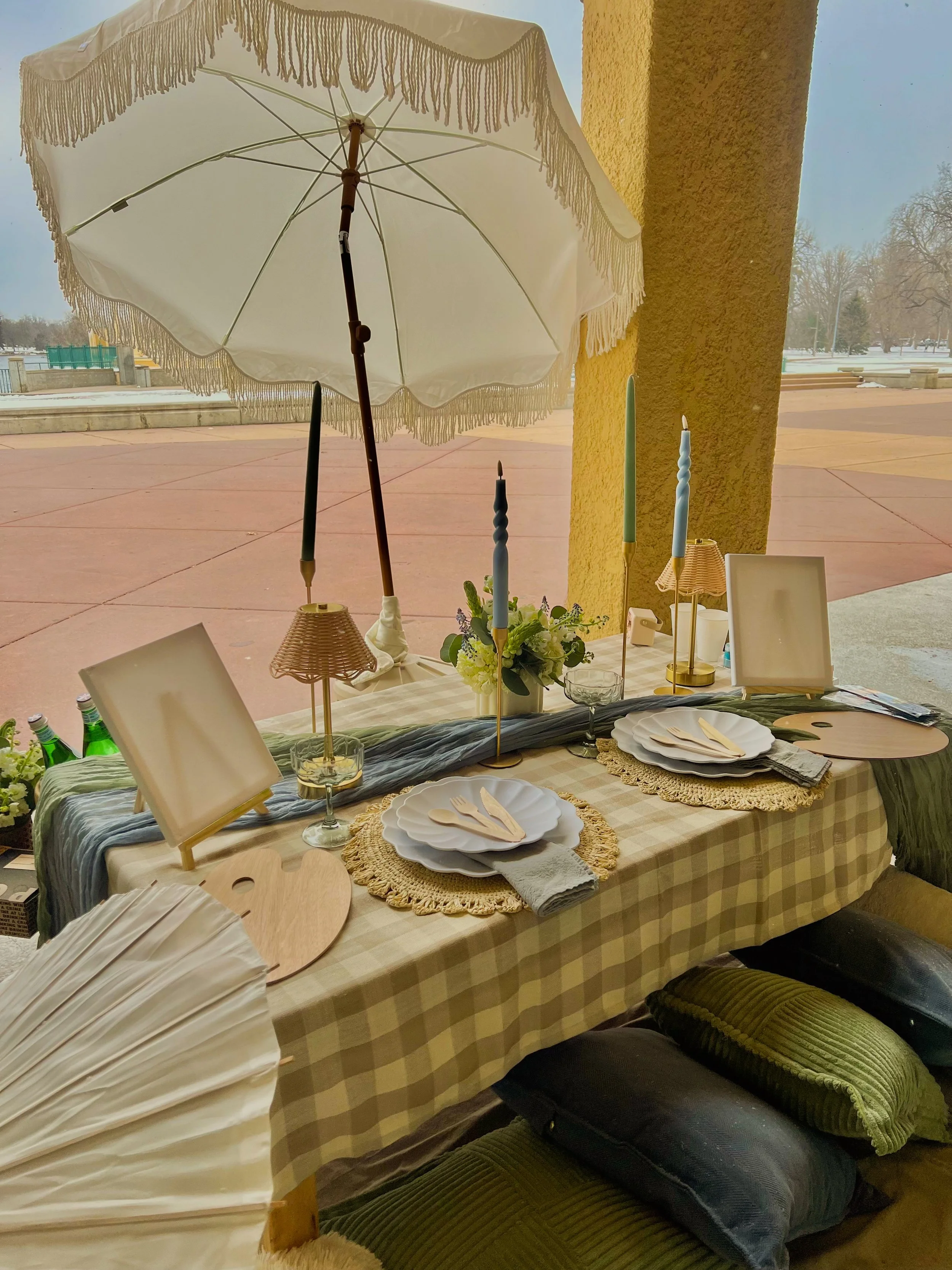 A decorated table with white plates, napkins, cutlery, green and black pillows, and a large white umbrella outside on a snowy day.