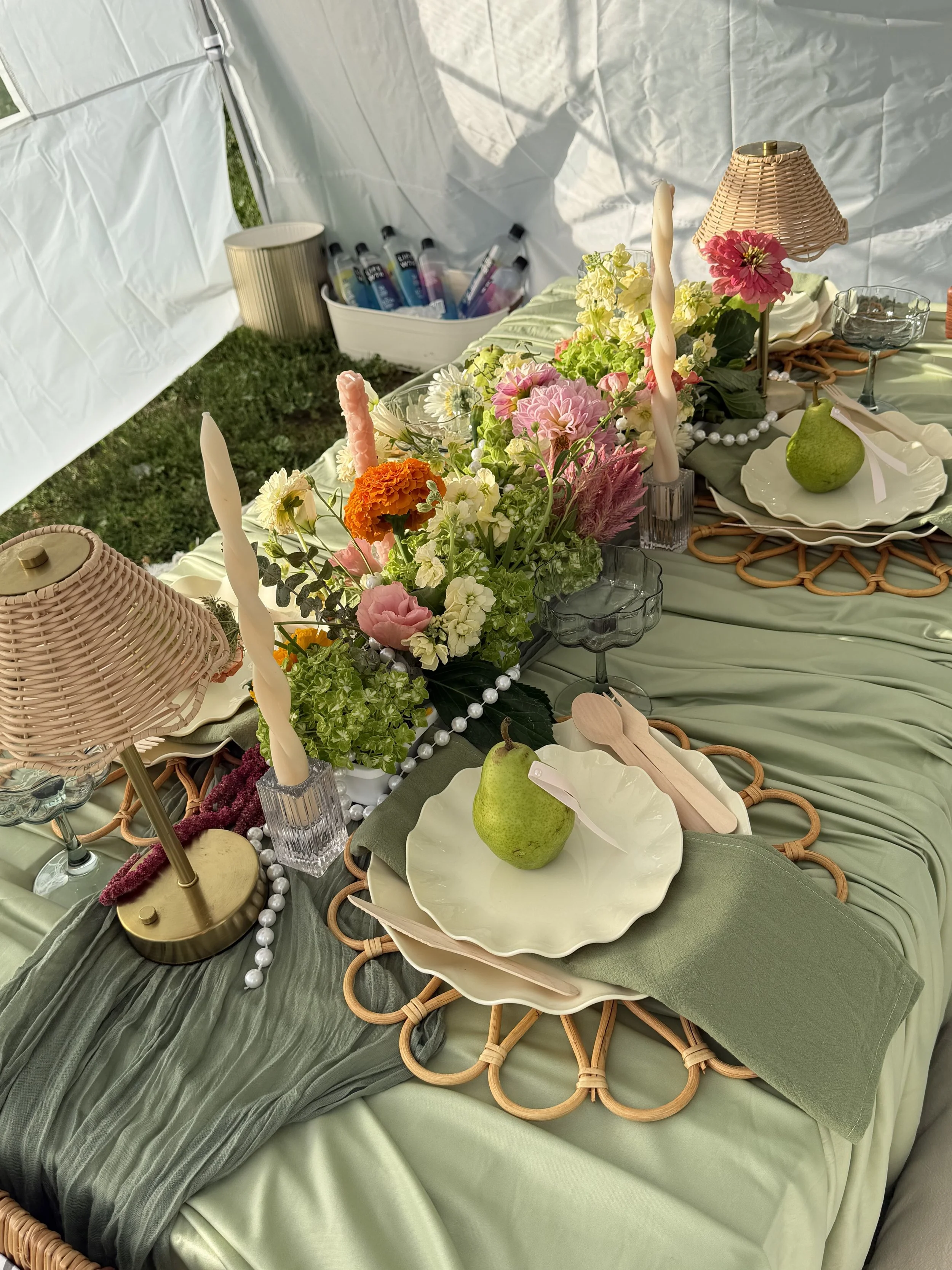 A picnic table set for a meal with a green tablecloth, floral centerpiece, white plates with pears, pink and white candles, wicker lampshades, glassware, and pearl decorations.