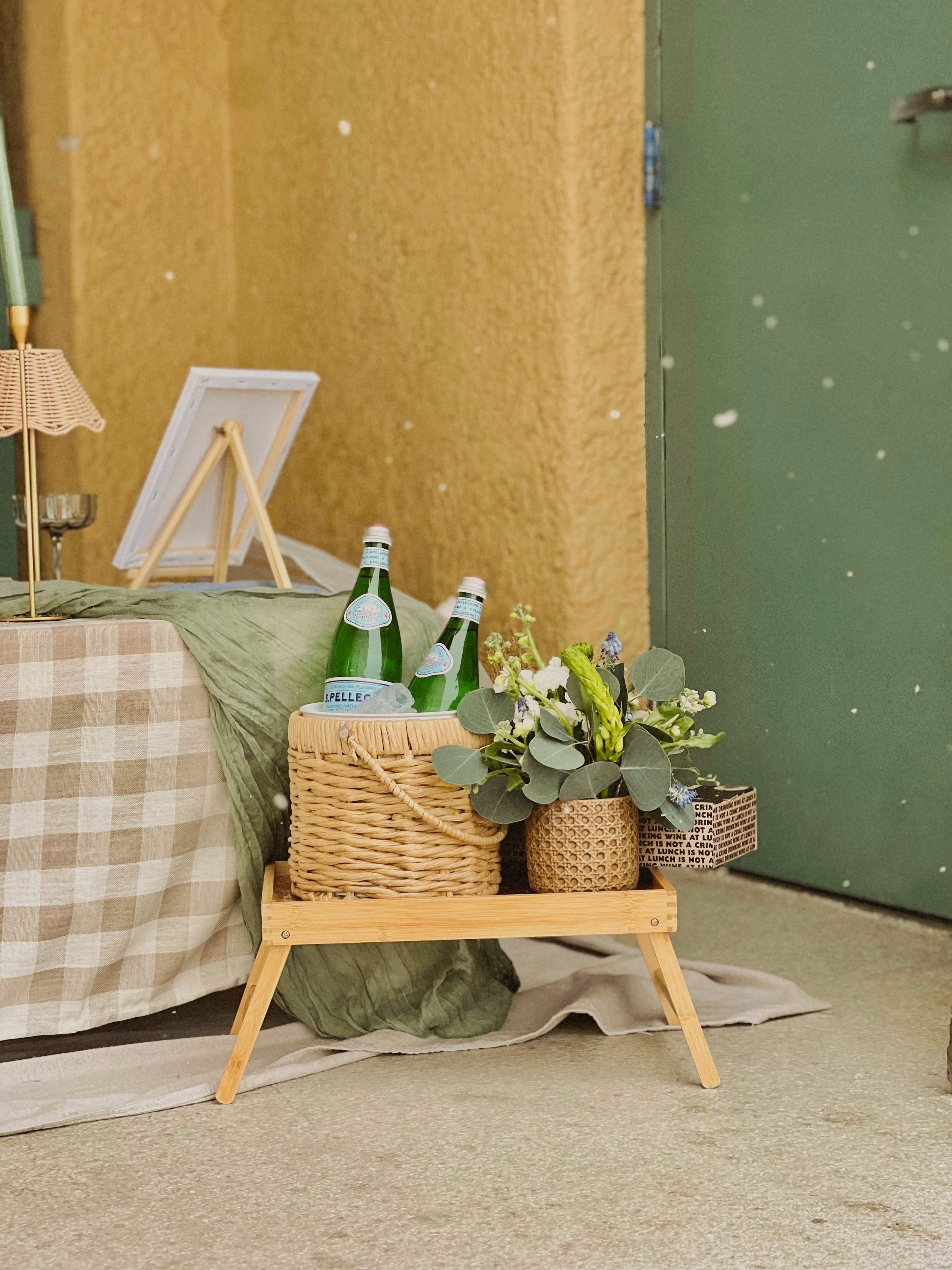 Close-up of a wooden tray on the floor holding a woven basket with green leaves and small flowers, two glass bottles of San Pellegrino sparkling water.