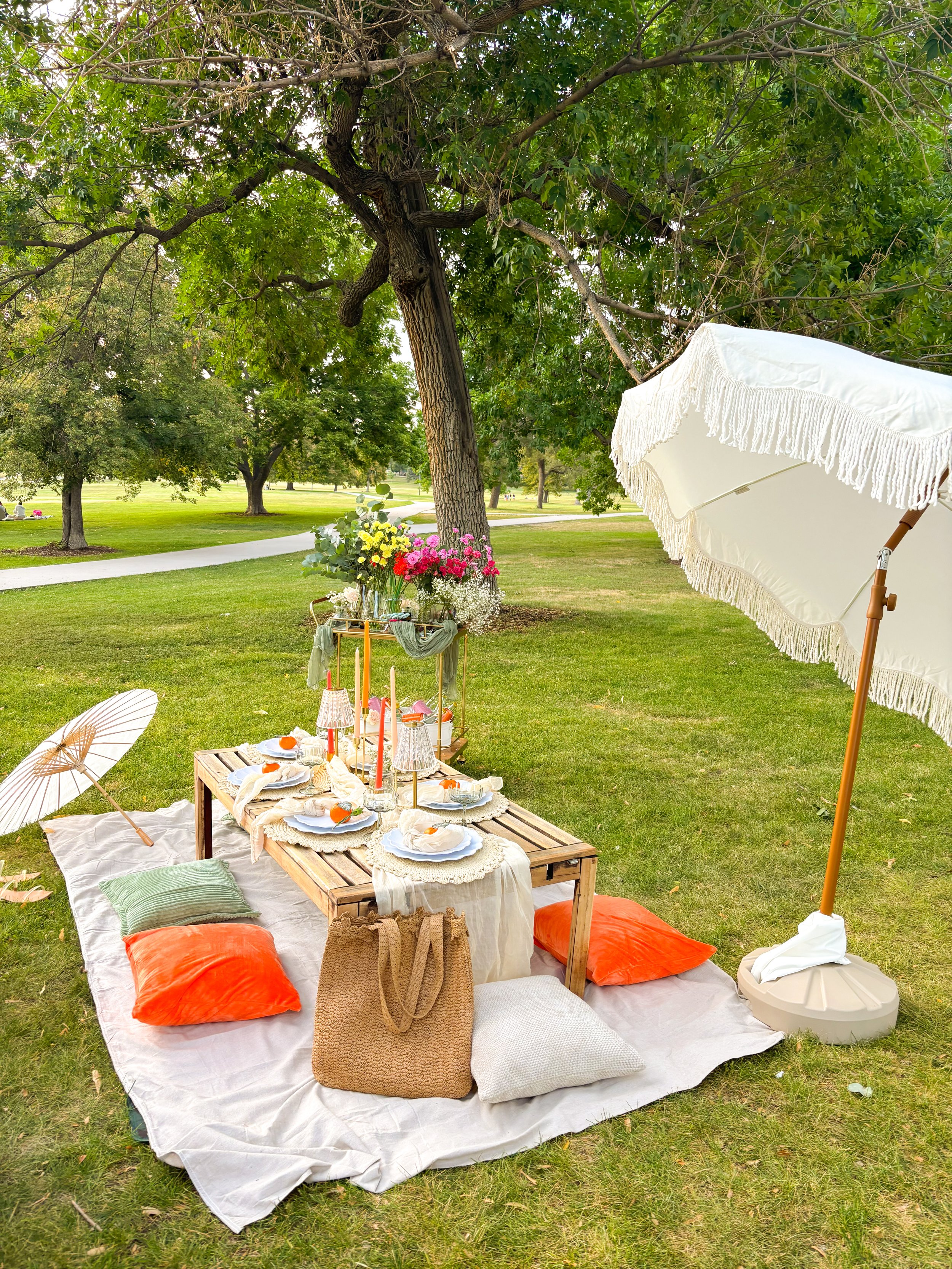 A picnic setup in a park with a low wooden table, sitting on a large white cloth. The table has plates, glasses, lit candles, and flowers. Orange cushions are on the ground nearby, along with a white parasol with fringes providing shade. There are tr