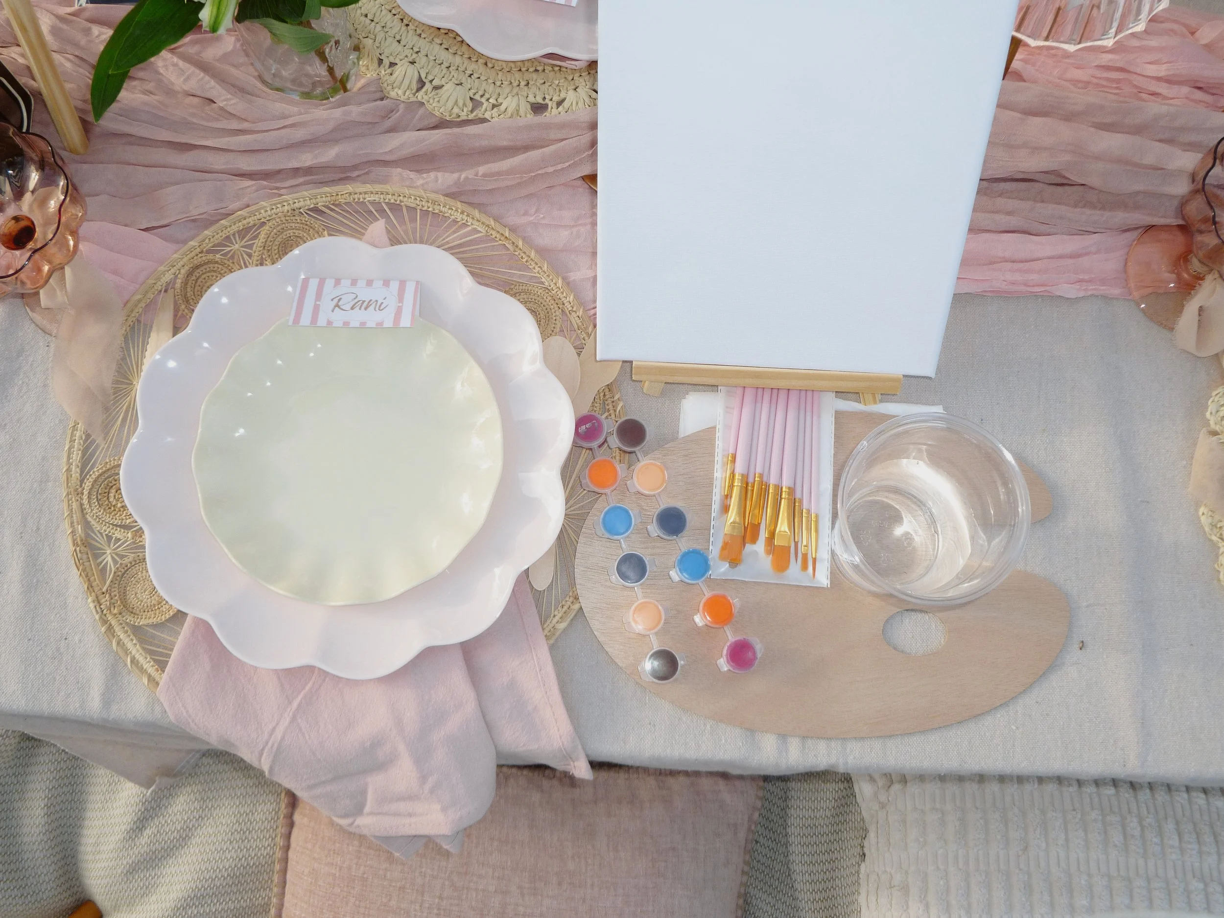 Decorative table setting with a white scalloped plate, small jars of colorful paint, glass of water, and a backdrop of pink fabric, green leaves, and candles.