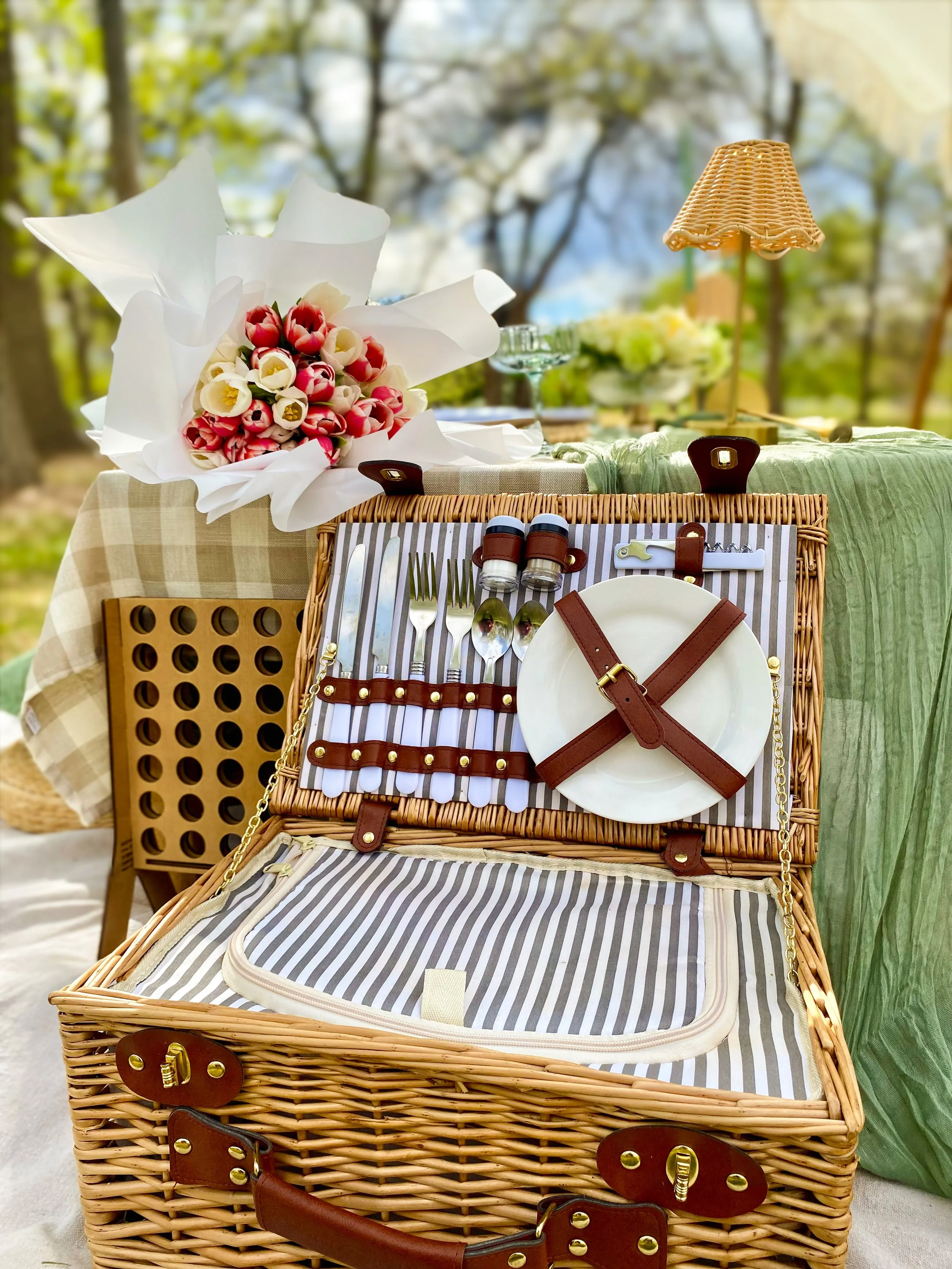 A picnic basket partly opened, containing plate, a utensil holder with knives, forks, and spoons. Behind the basket, there is a large bouquet of pink and white flowers wrapped in white paper. To the right, there's a small wicker lamp with a shade, an