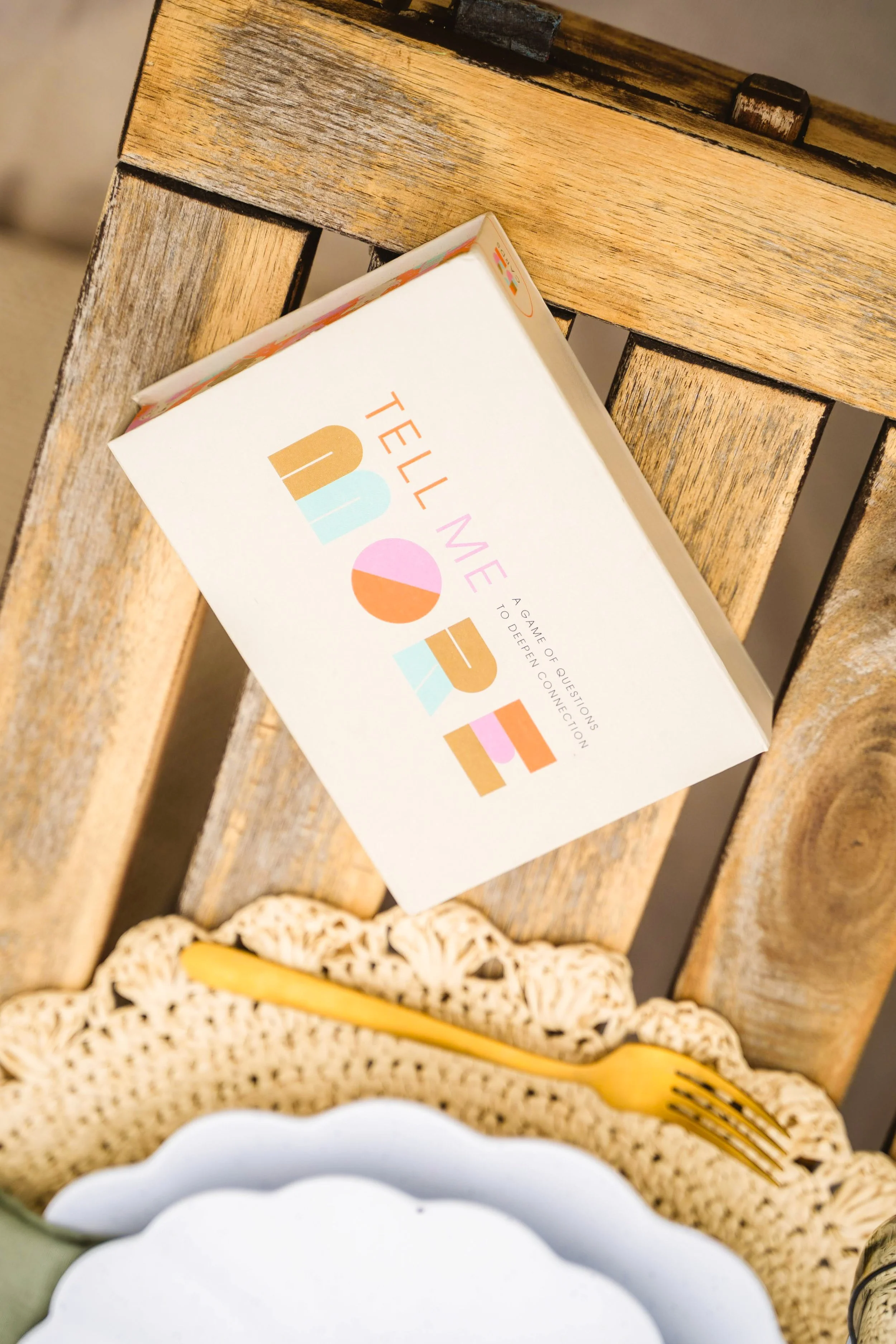Close-up of a wooden dinner table with a white box of colorful text titled 'Tell Me More,' a gold fork, and a ornate lace placemat.