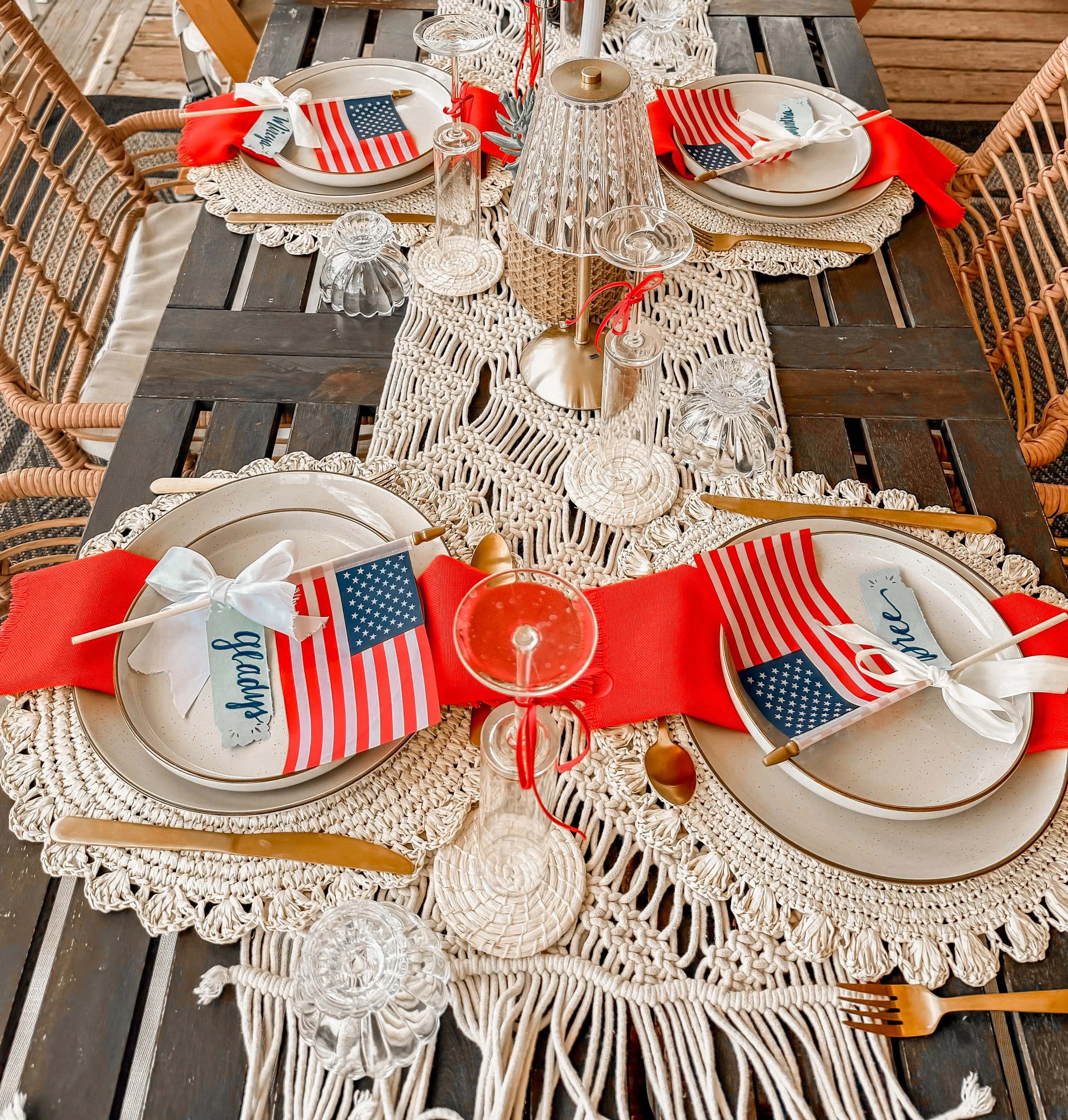 Table set for an American holiday celebration. It features plates with napkins, small American flags, glasses, and decorative elements like a woven table runner and a lamp, all arranged on a wooden table with wicker chairs around it.