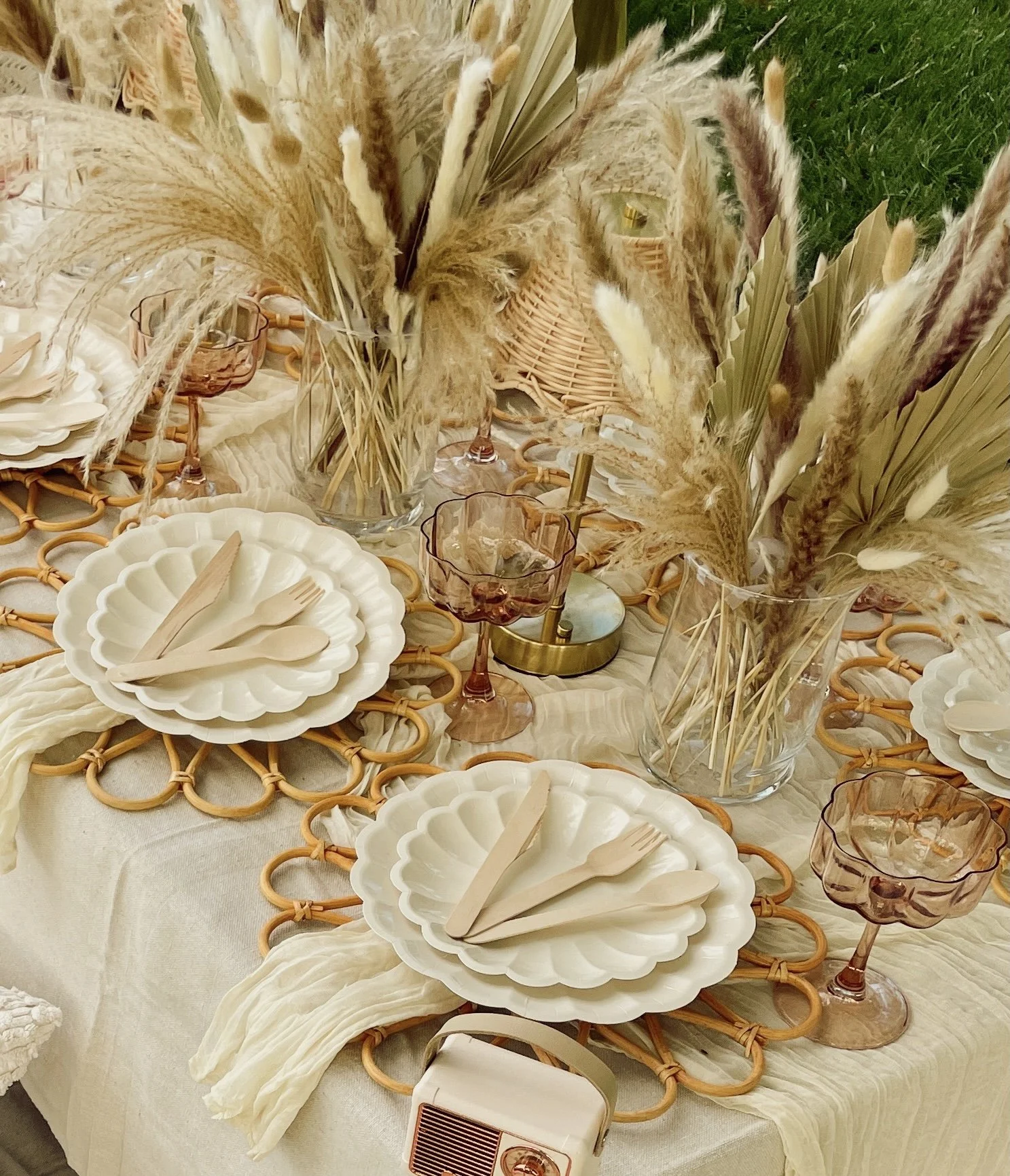 Table set with white scalloped plates, wooden cutlery, pink glassware, and tall vases holding dried pampas grass and other dried plants.