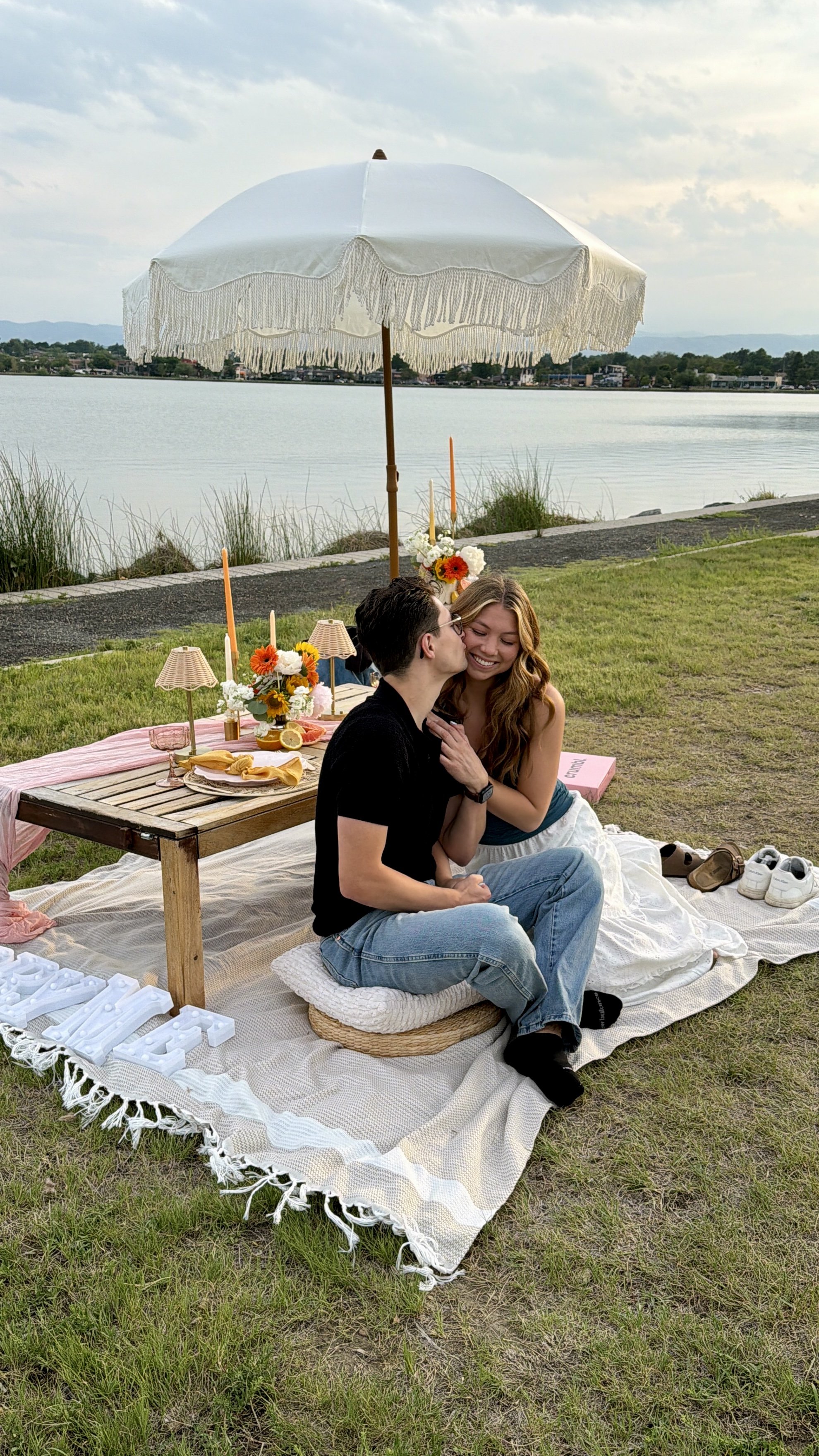 A couple sitting on a blanket by a lake having a luxury picnic with a table decorated with flowers and candles. A white parasol shades them.