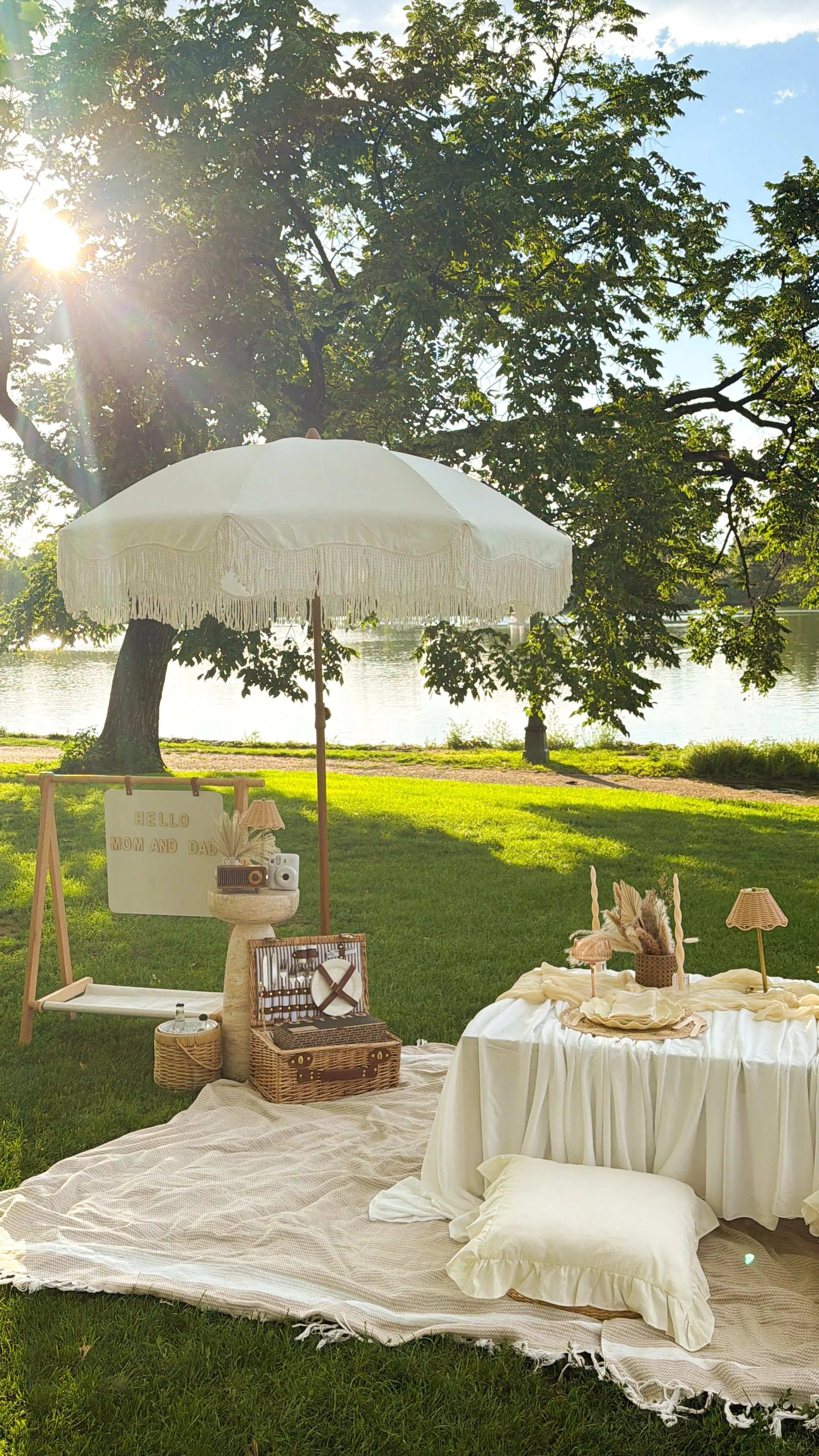Outdoor luxury picnic with a white umbrella, a white table with decor, and a sign that reads 'Hello Mom and Dad' near a lake, with green grass, trees, and sunlight.